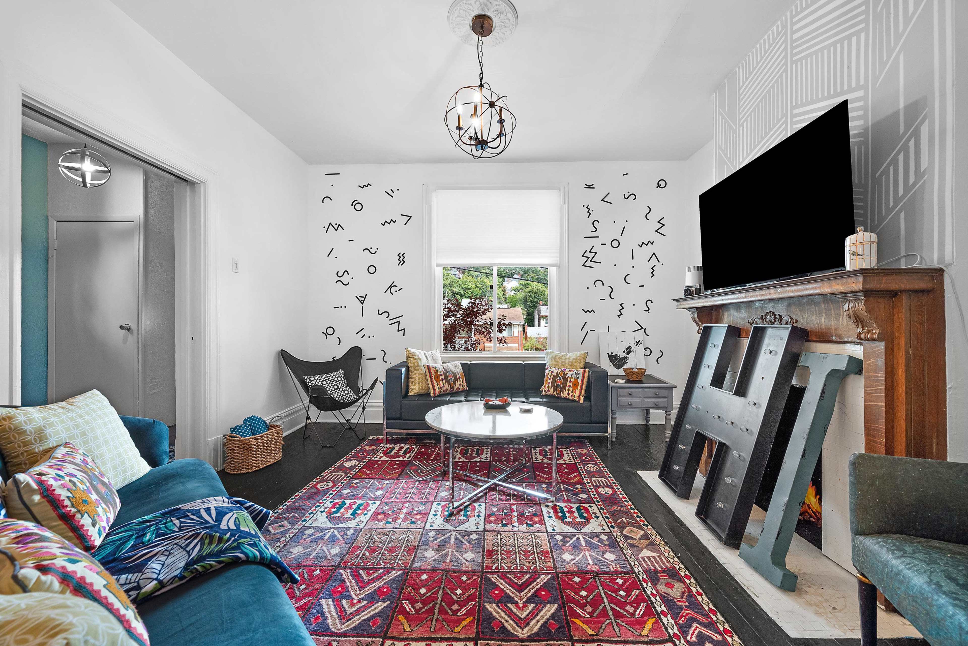 The living room features a patterned rug, two sofas, a circular coffee table, and a decorative fireplace with a large television mounted above it.
