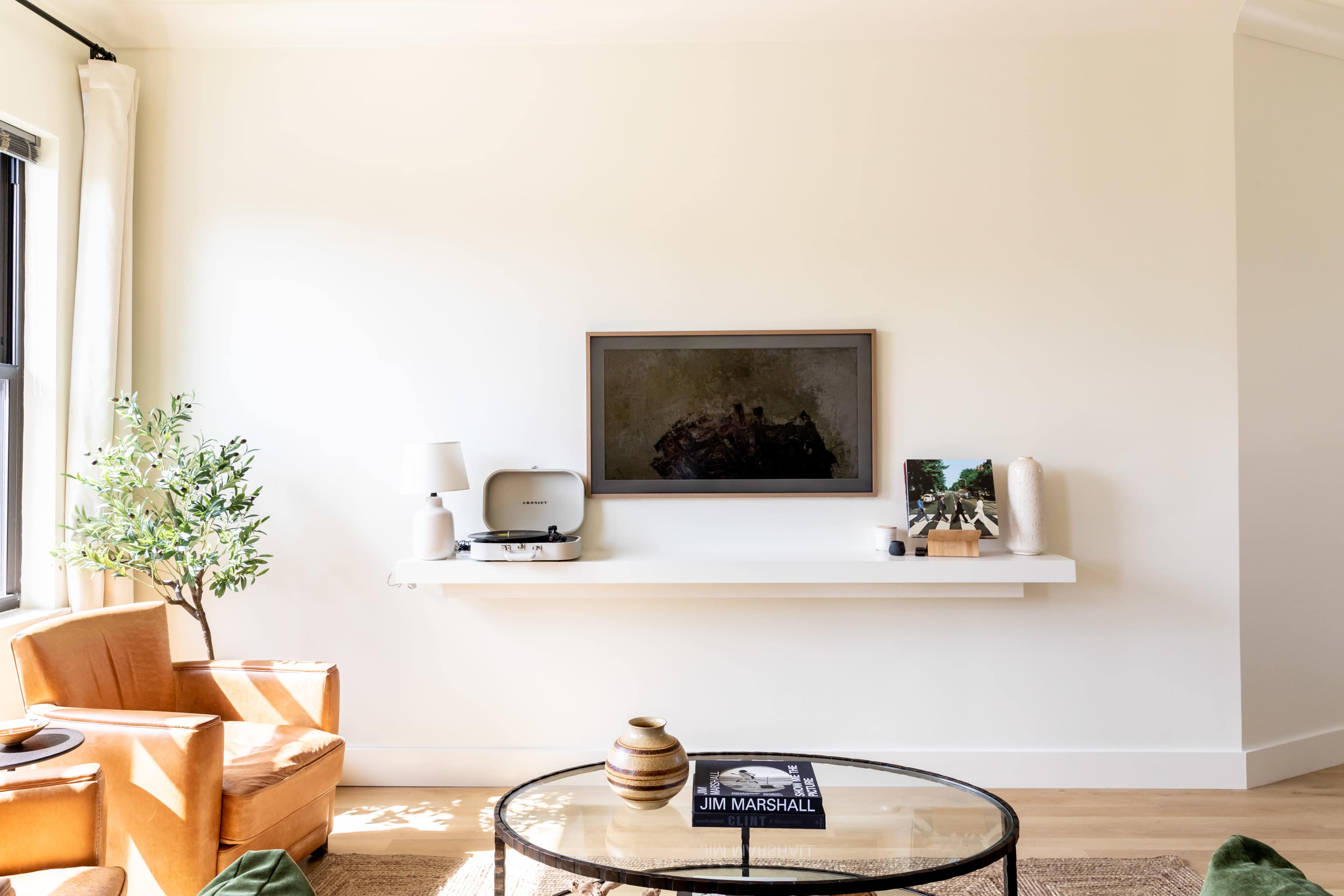 A cozy living room features an orange armchair, a glass coffee table with a book, a small plant, and a wall-mounted shelf with a record player and decorative items.