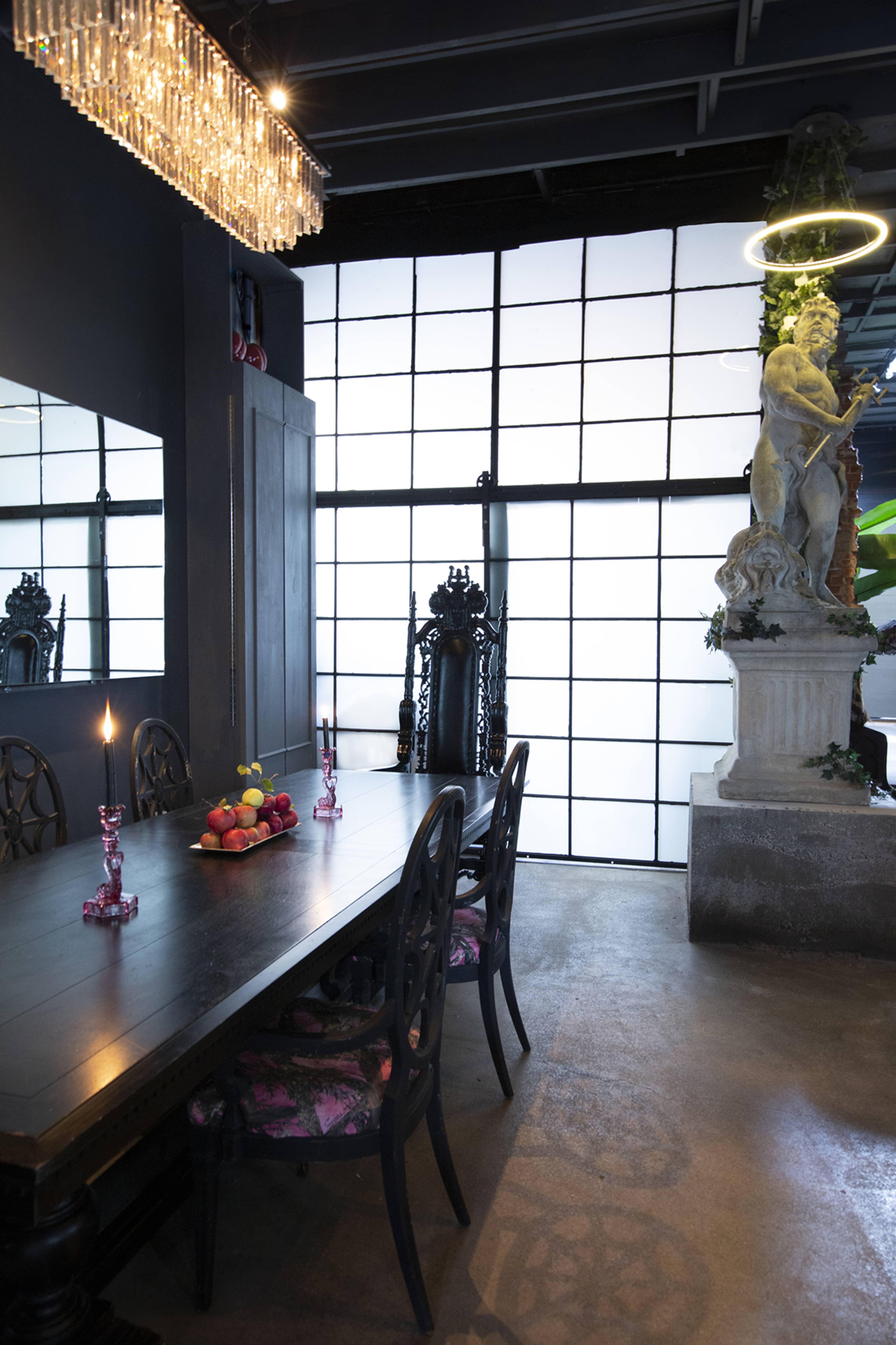 The image shows a dining area with a long black table surrounded by ornate chairs, a centerpiece of fruit, and a large window creating a backdrop with a statue nearby.