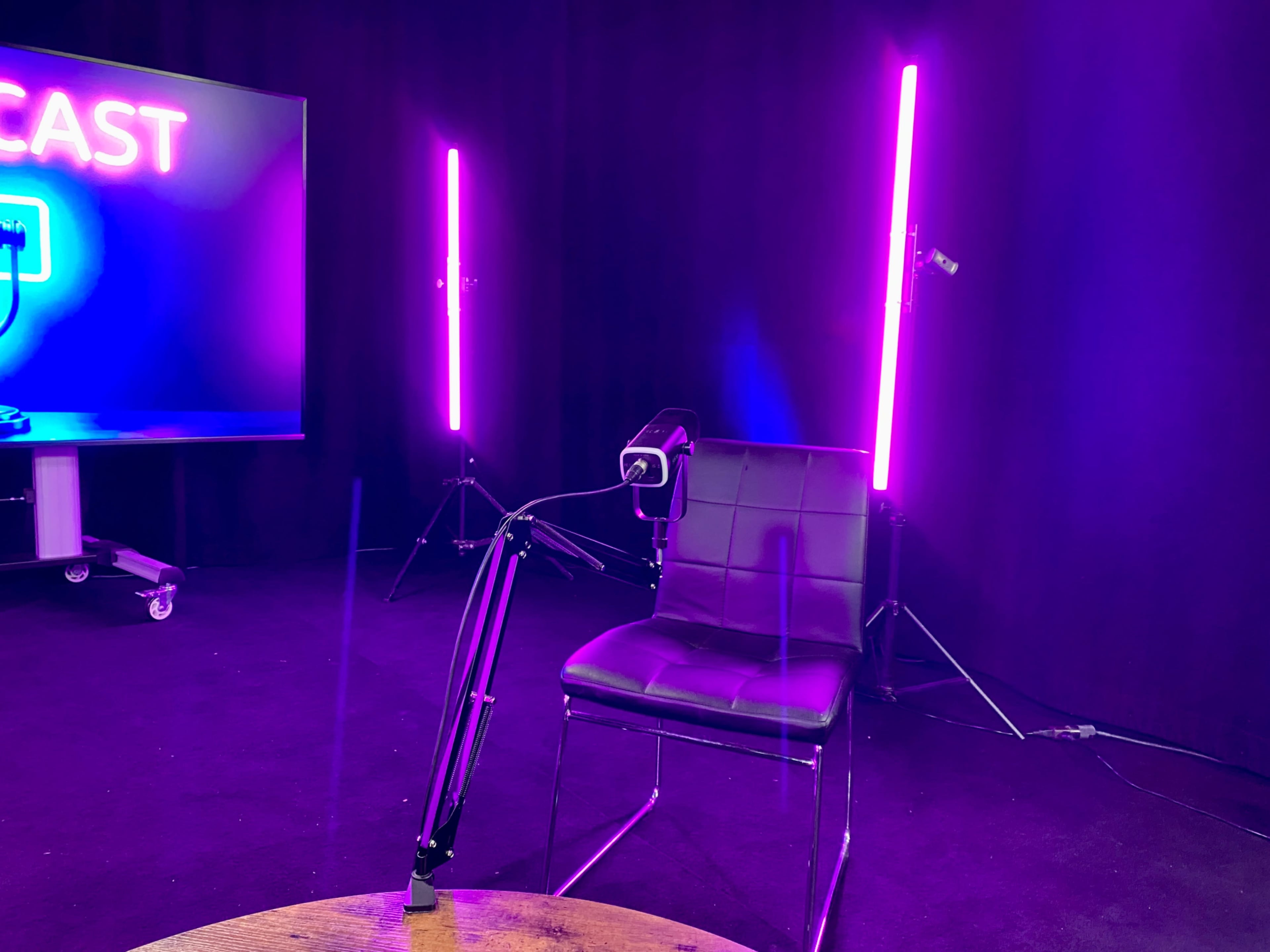 A black chair is positioned in front of a display screen with the word "PODCAST" illuminated in neon lights, flanked by two vertical purple lights.