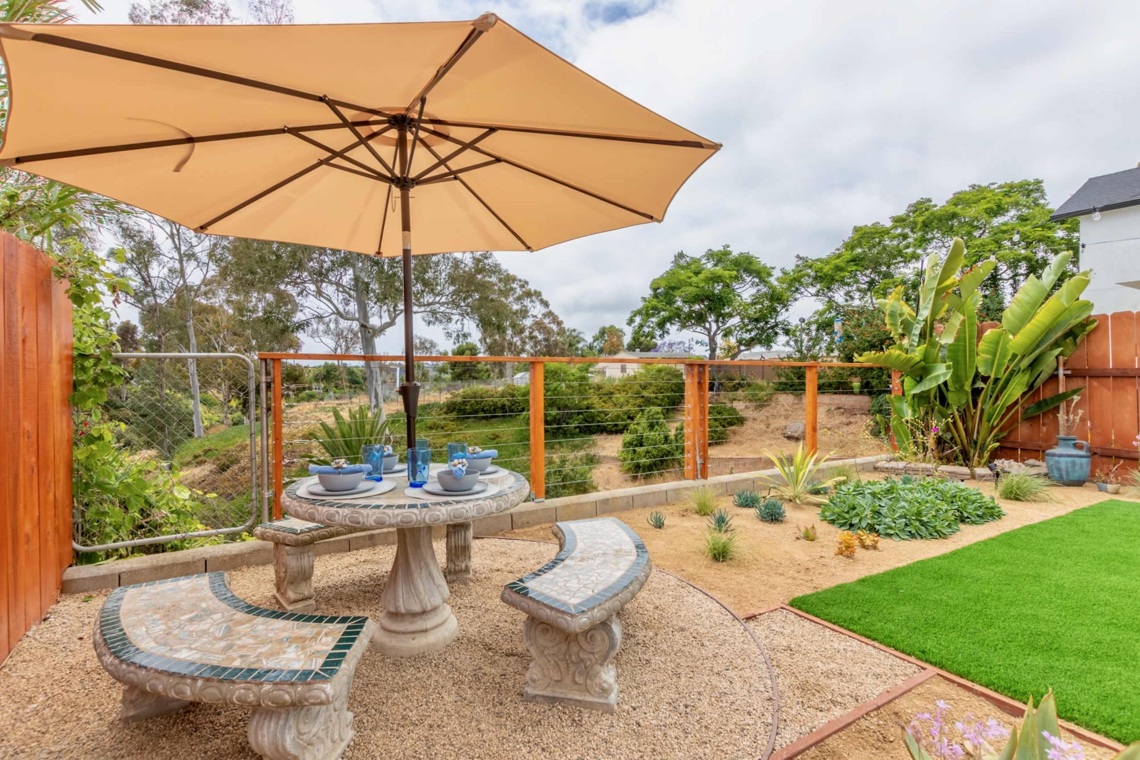 A stone table with matching curved benches is set under a large umbrella in a landscaped yard featuring greenery and decorative plants.