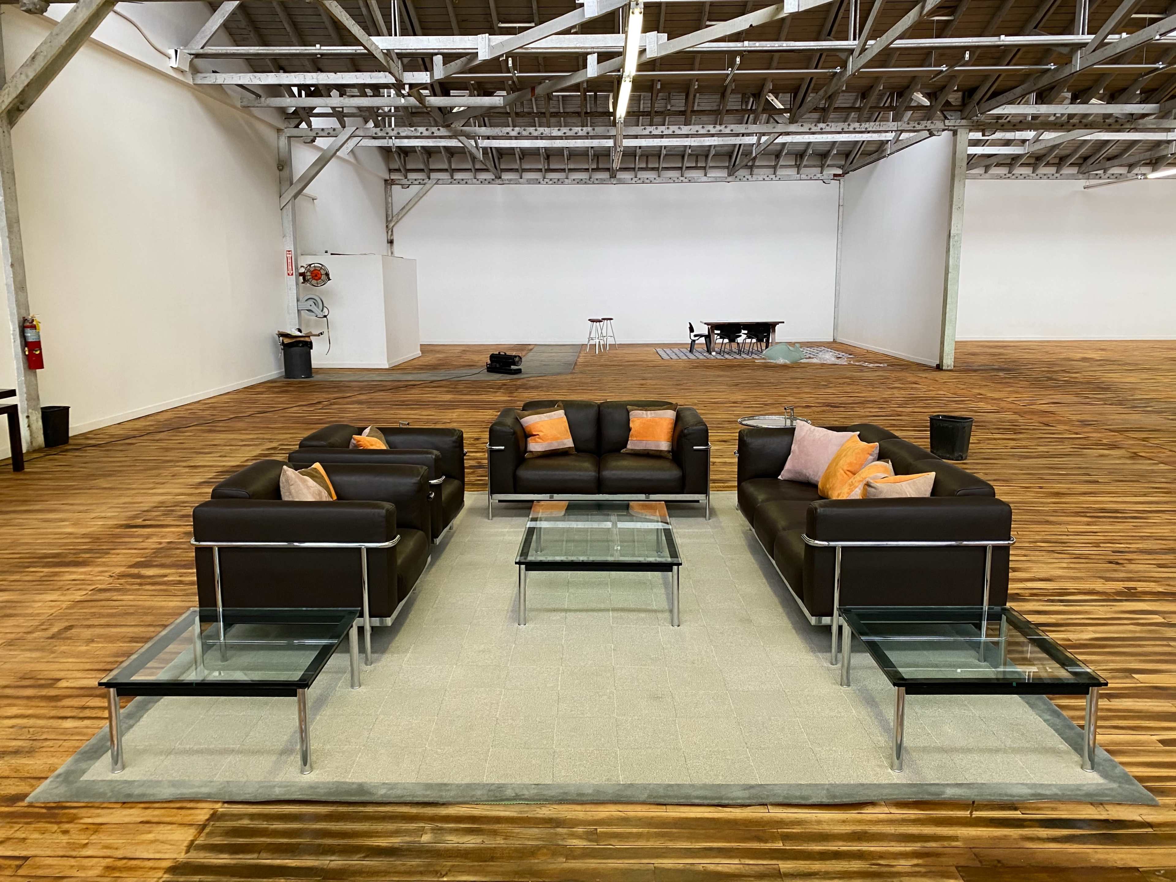 A set of black leather couches arranged in a square around a glass coffee table on a gray rug in an empty, spacious warehouse interior.
