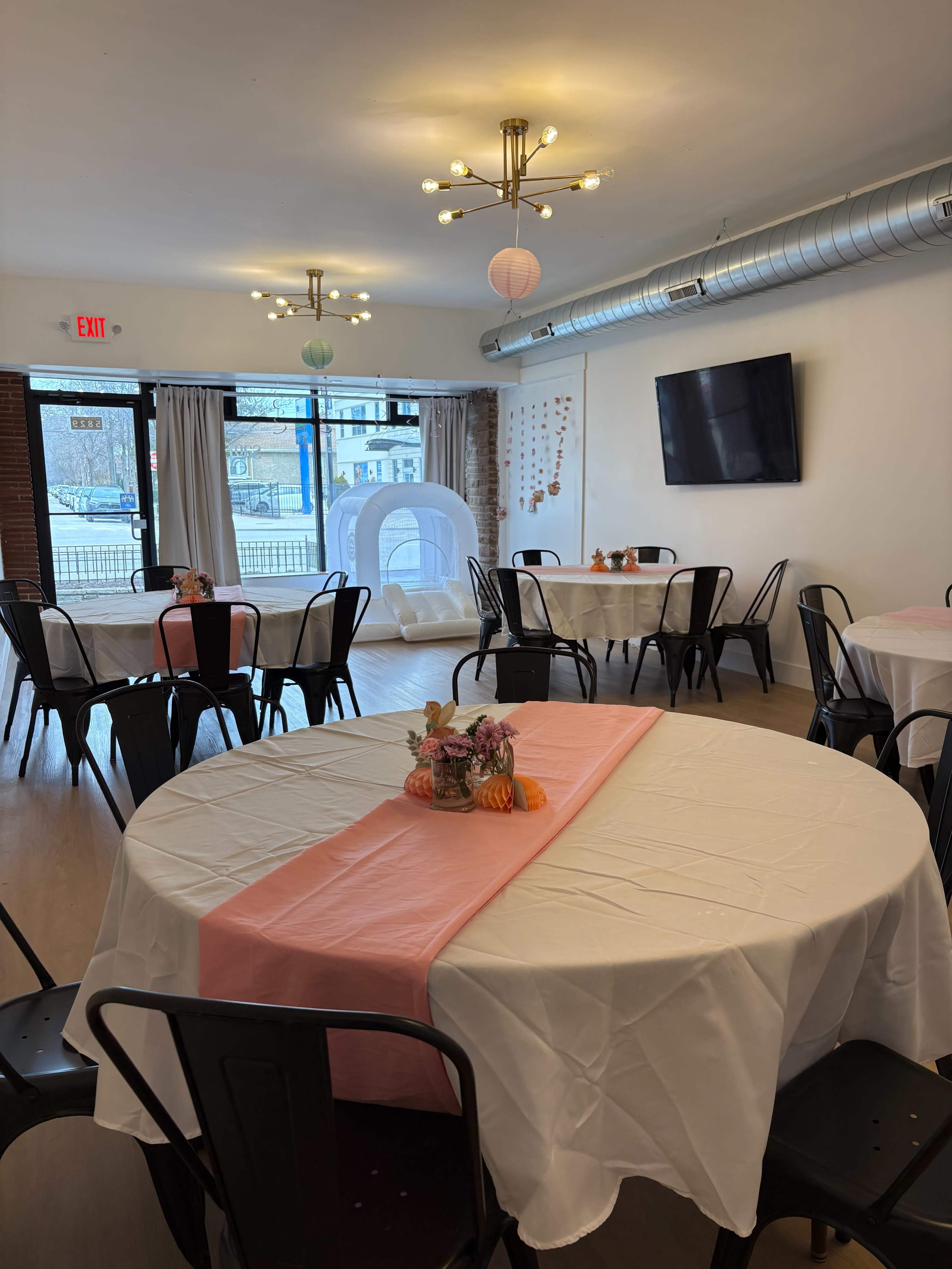 The image shows an indoor event space with round tables covered in white tablecloths, each adorned with a centerpiece, and pastel decorations on the walls.