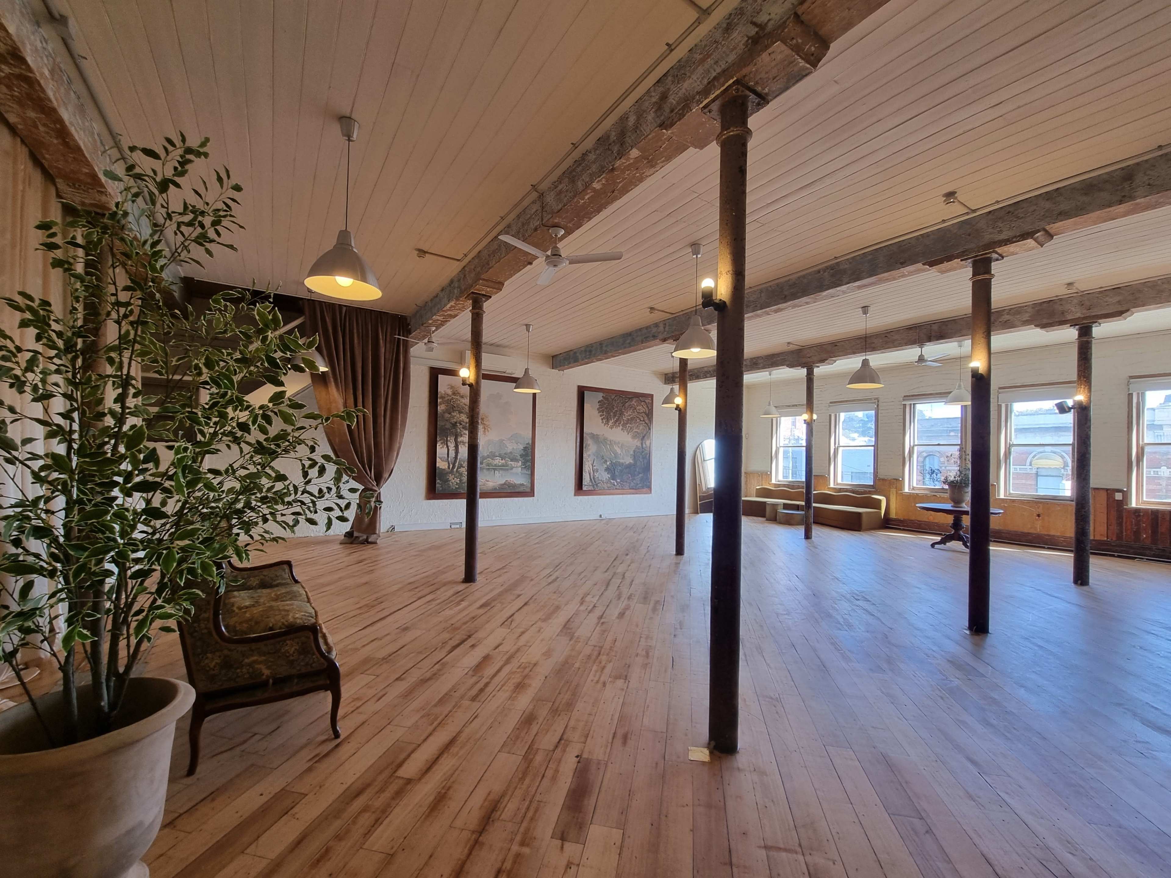 The image shows a spacious, well-lit room with wooden floors, high ceilings, and large windows, featuring artwork on the walls and a few pieces of furniture.