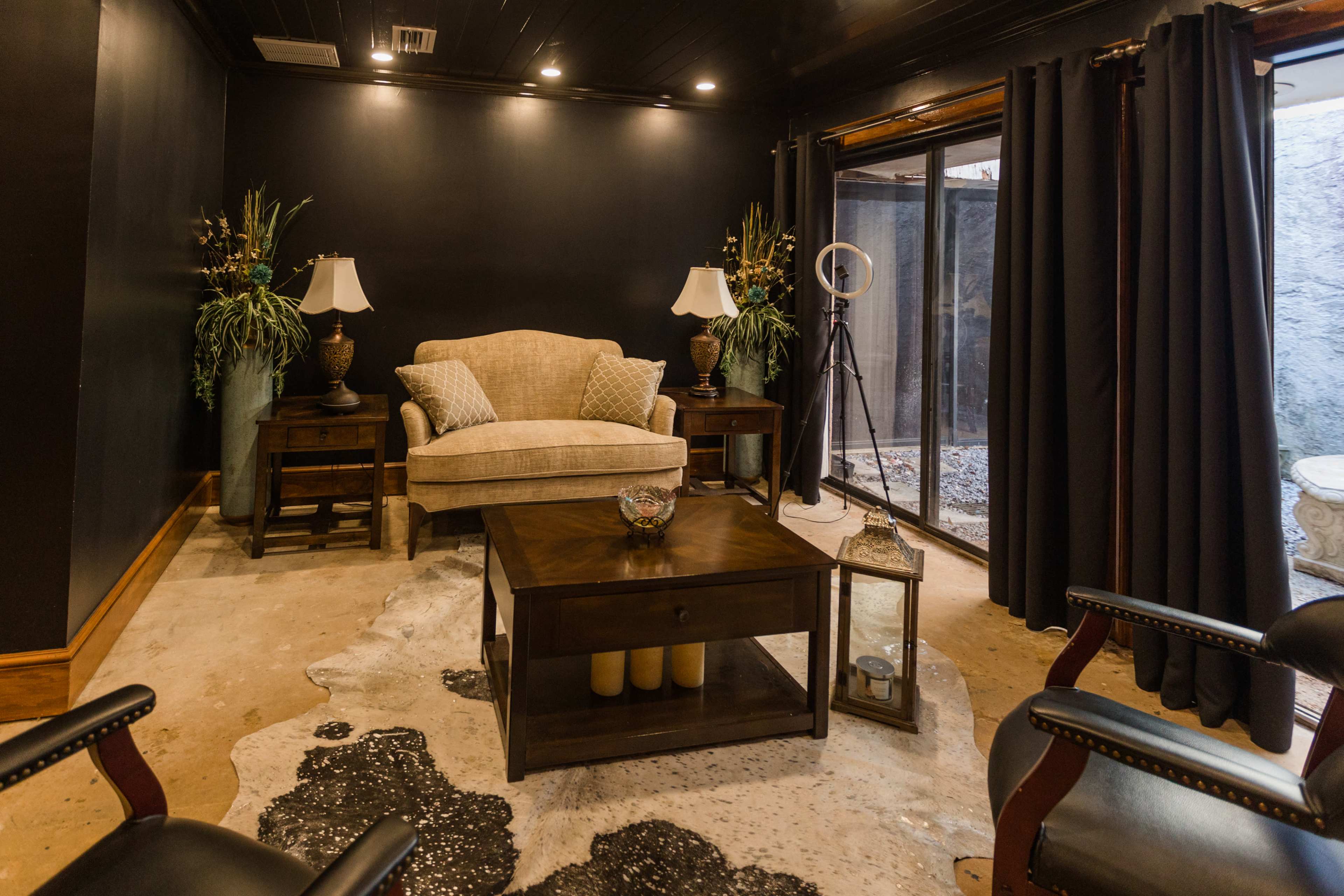 A cozy room with a beige couch flanked by table lamps, a dark wooden coffee table in the center, and a large window with dark curtains revealing an outdoor view.