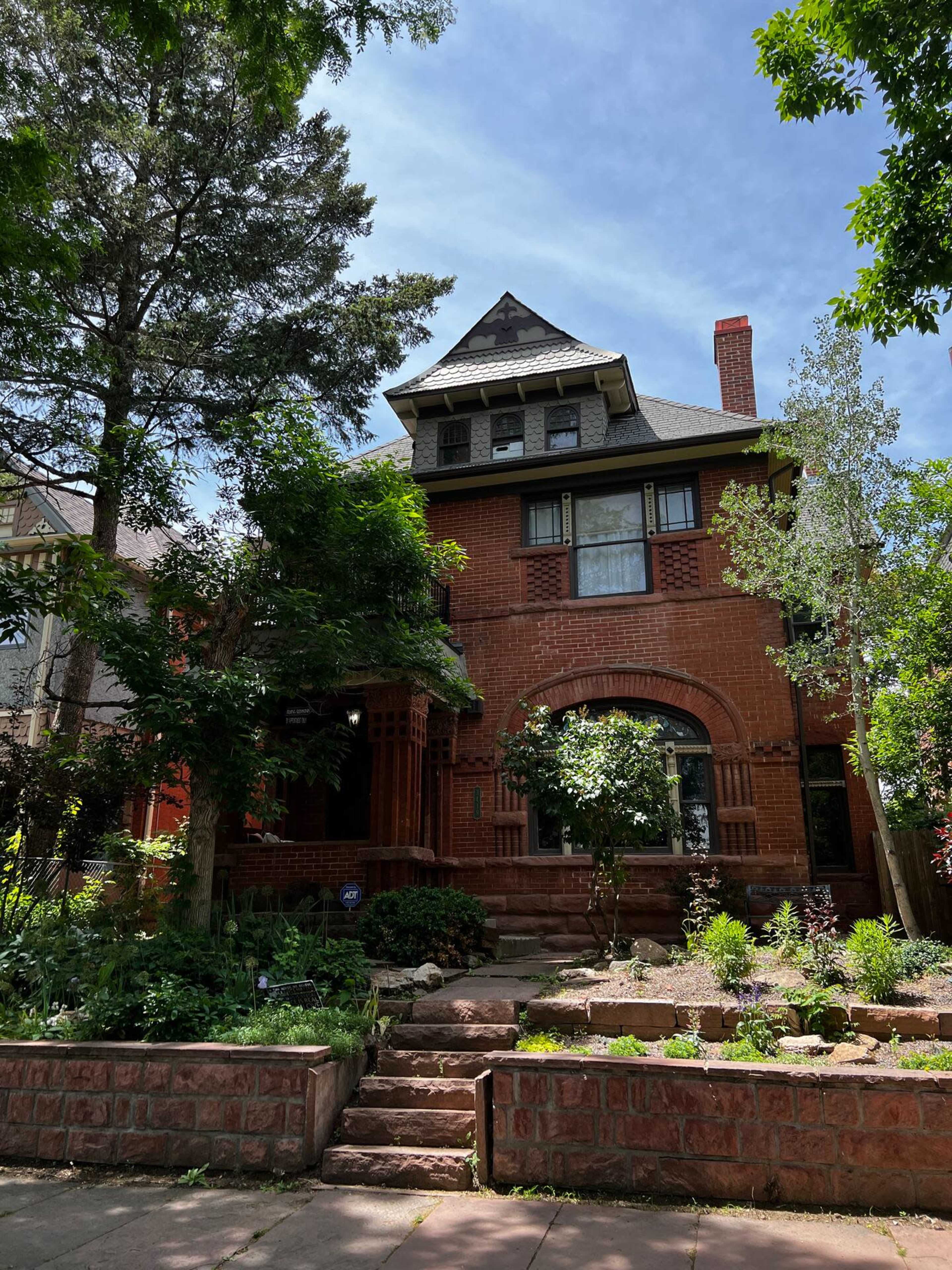 A three-story red brick house with a steeply pitched roof and a front garden featuring various plants and trees.