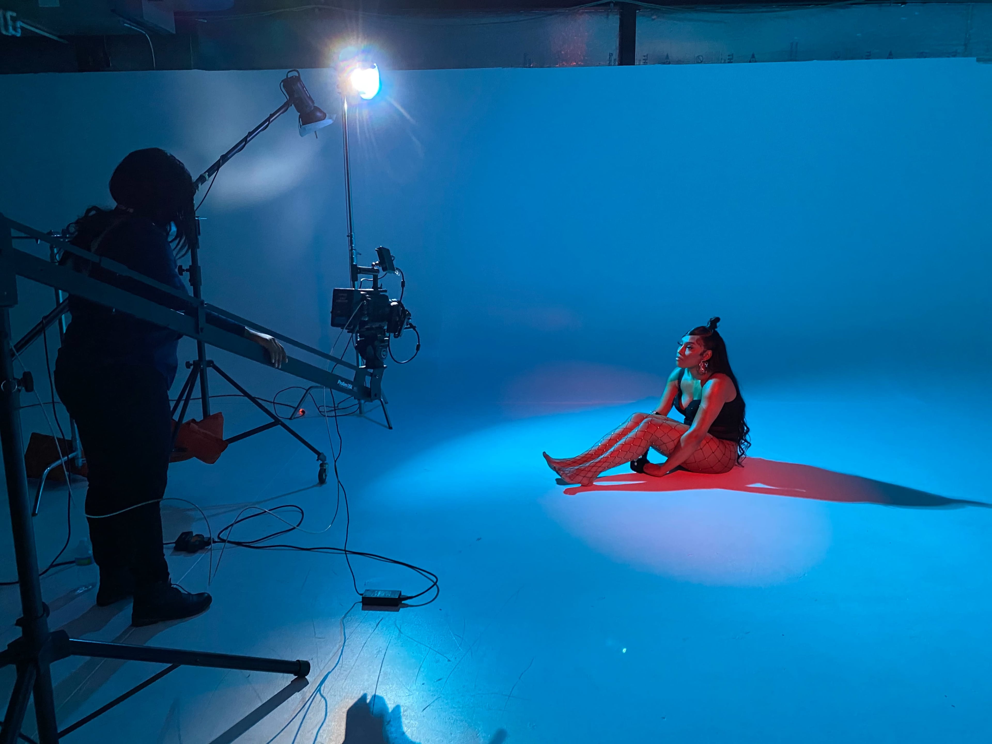 A model sits on the floor in a studio, illuminated by blue and white lights, while a camerawoman operates equipment nearby.