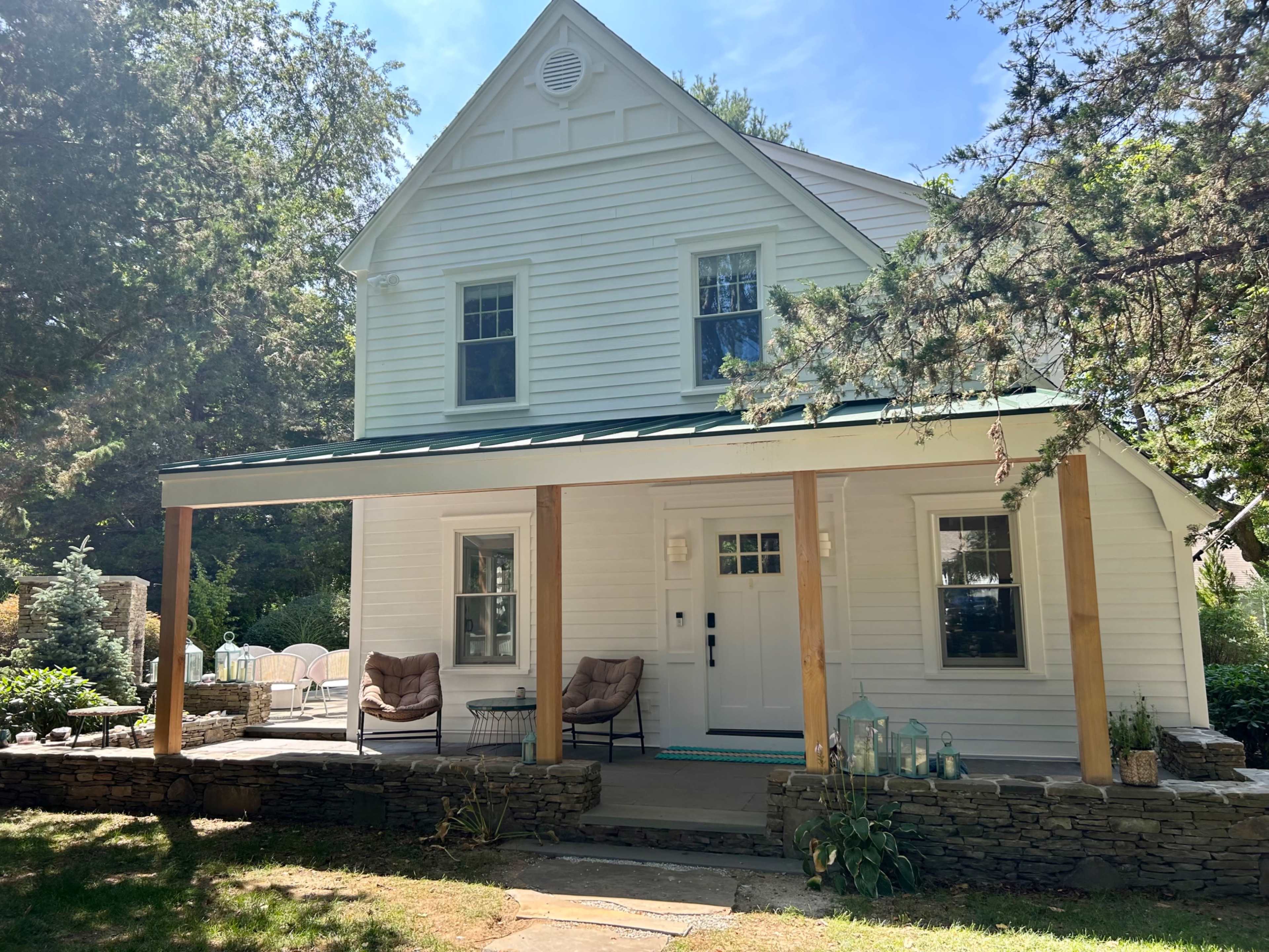 A two-story white house with a green roof features a front porch supported by wooden pillars.