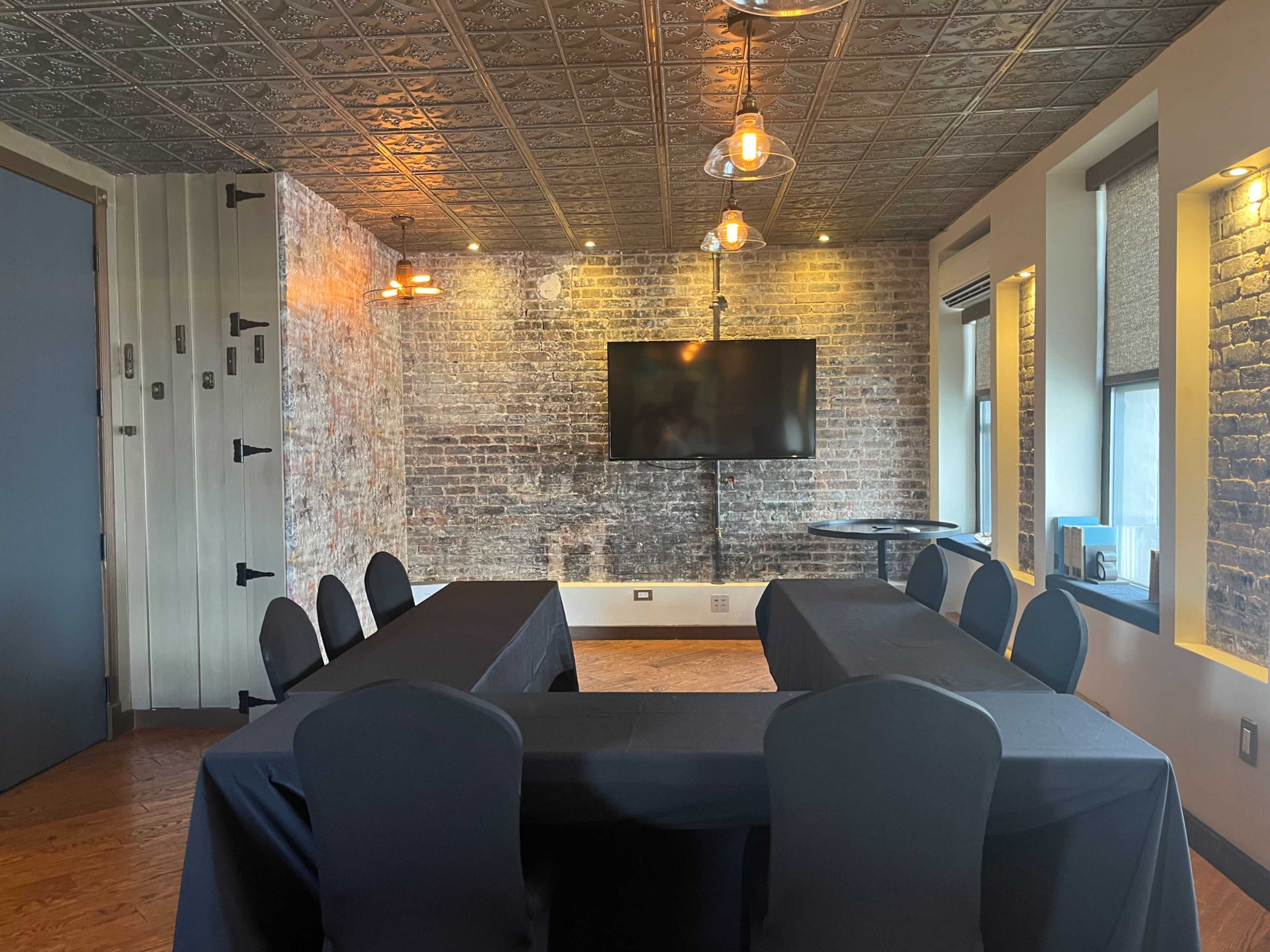 The image shows a conference room with a rectangular arrangement of tables covered in black cloth, exposed brick walls, and a large screen mounted on one wall.