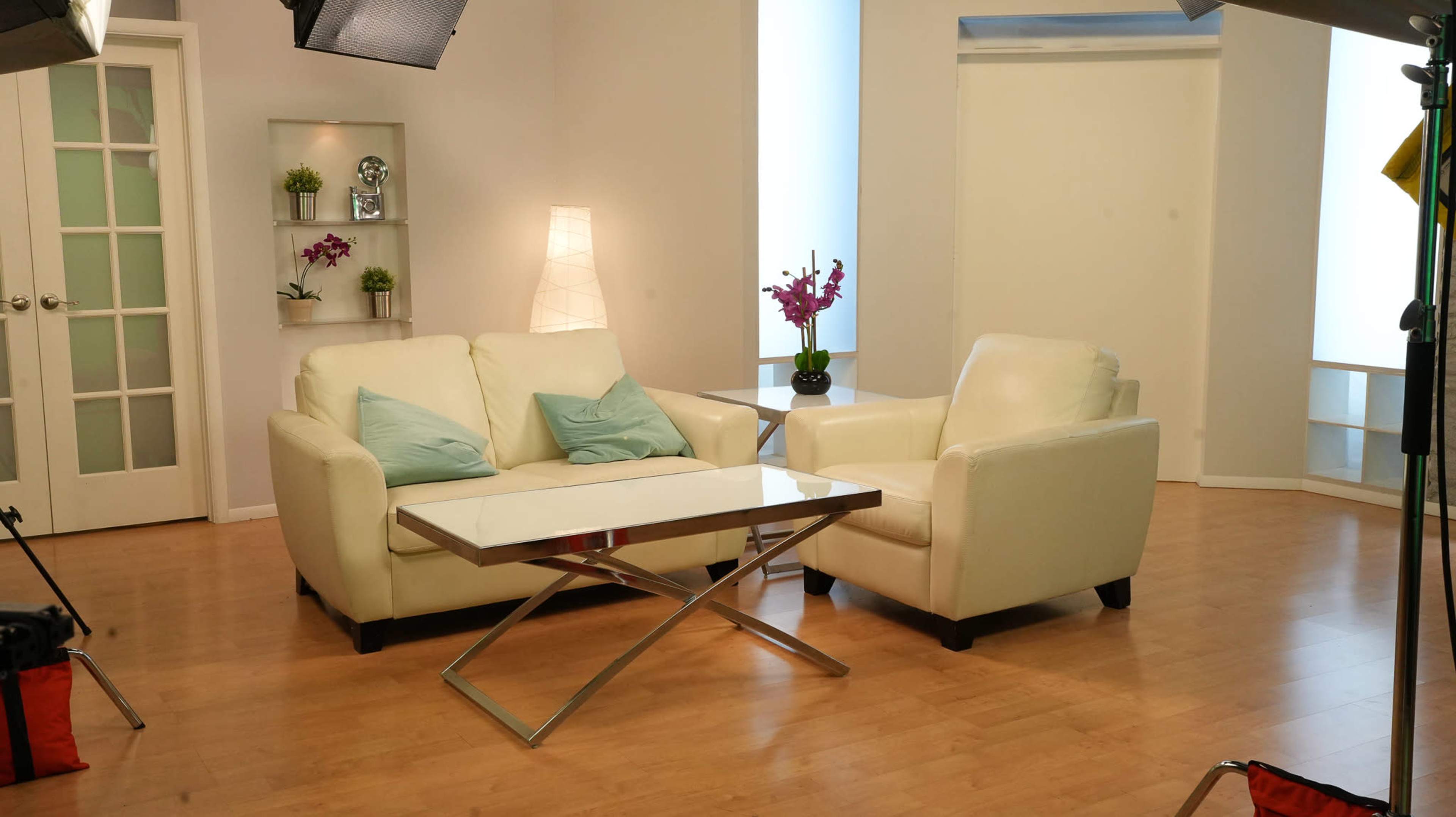 A modern living room scene features a pair of white leather sofas, a glass-top coffee table, and decorative plants, all set against light-colored walls and wooden flooring.