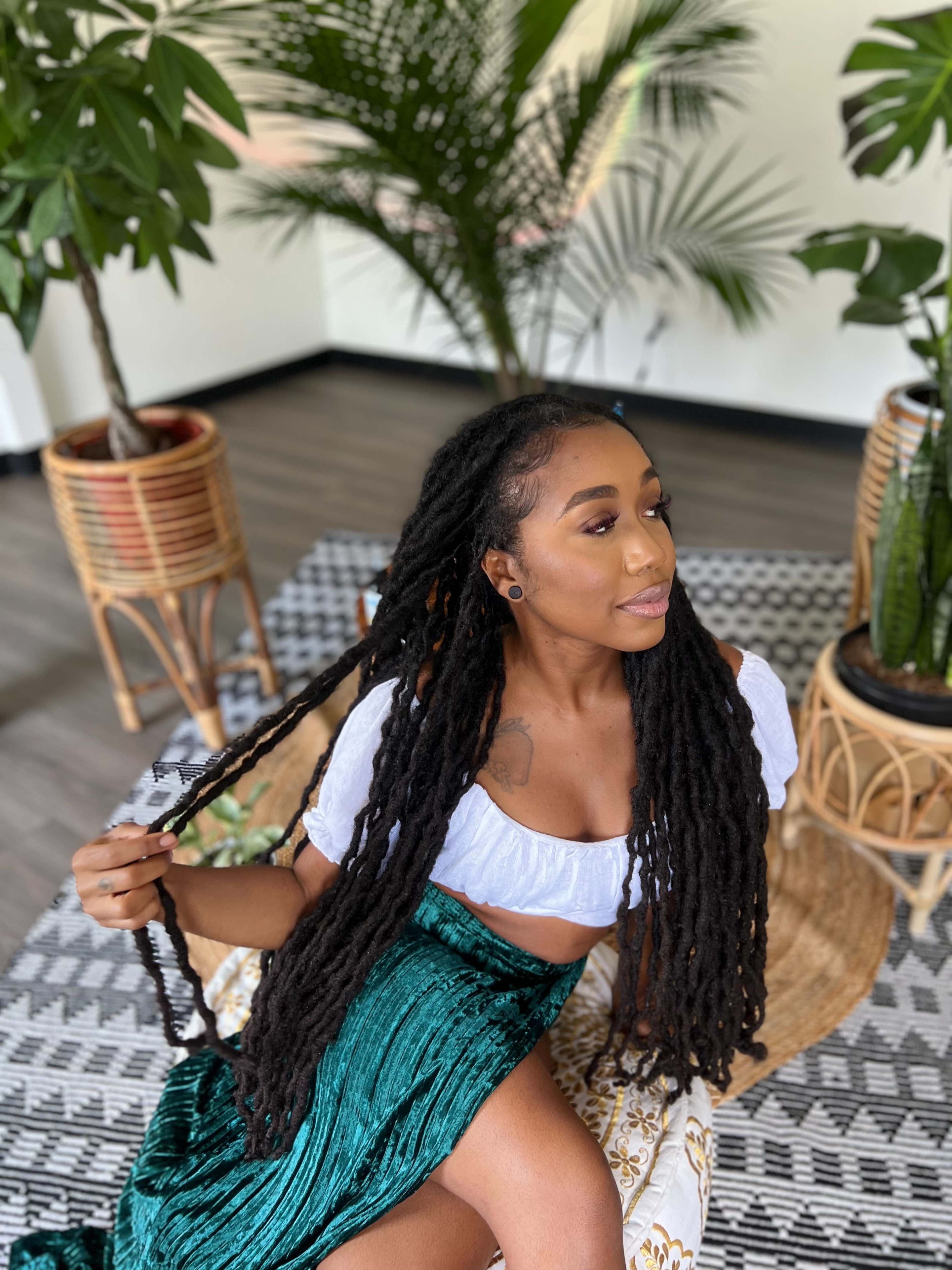 A woman with long, braided hair sits on a patterned rug surrounded by potted plants.