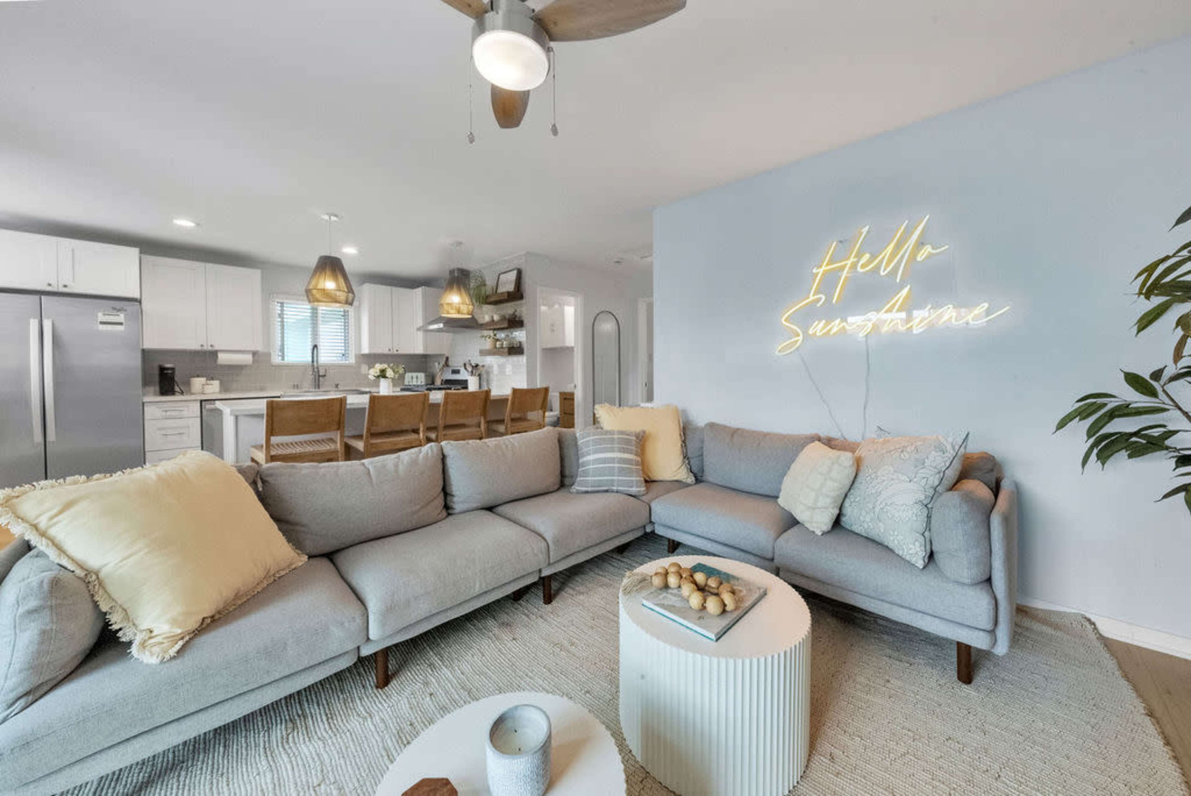The image shows a modern living room with a gray sectional sofa, a round coffee table, and a kitchen area visible in the background.