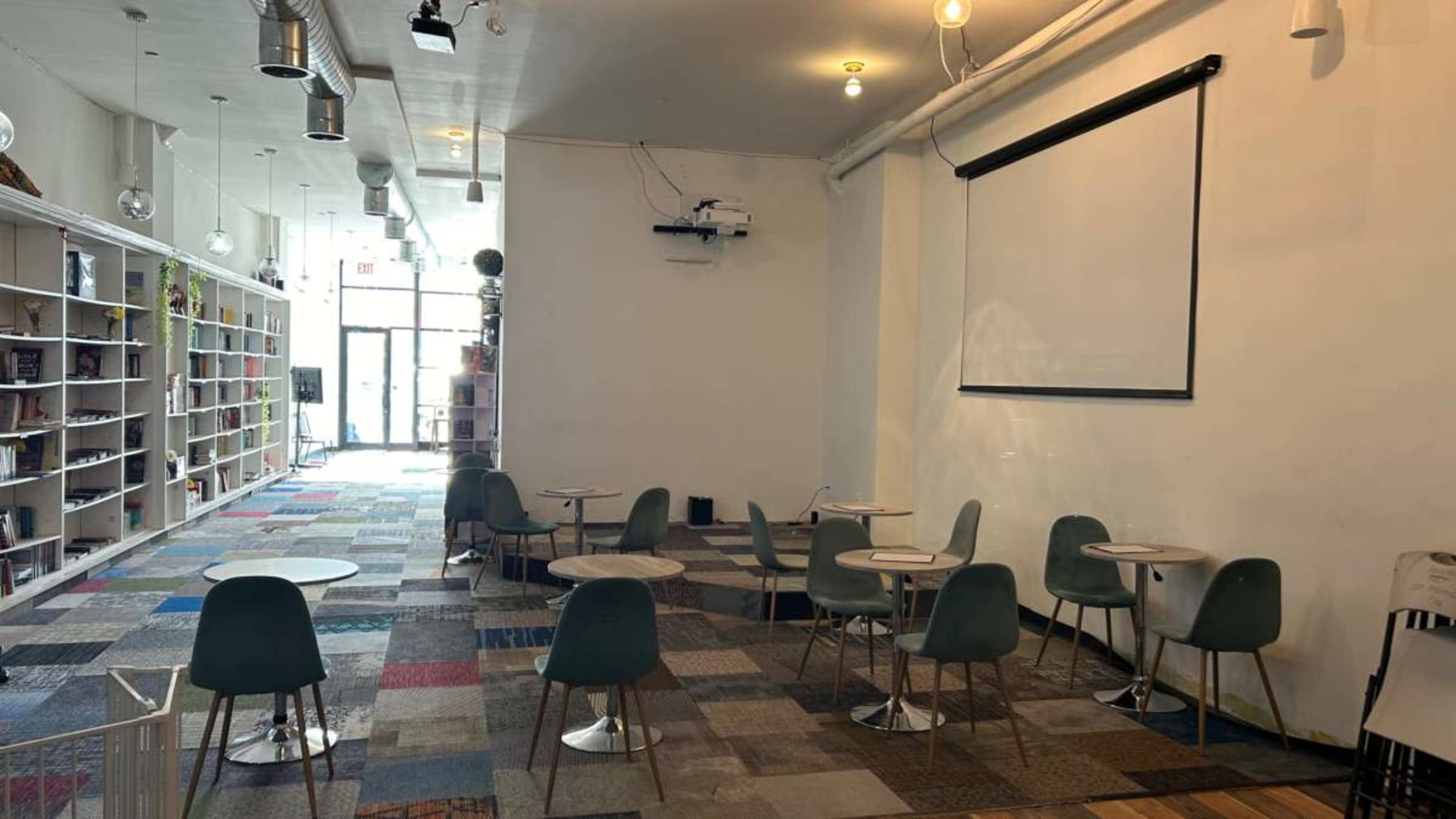 A spacious, minimalist café with several small tables and green chairs, a projector screen on the wall, and a bookshelf along one side.