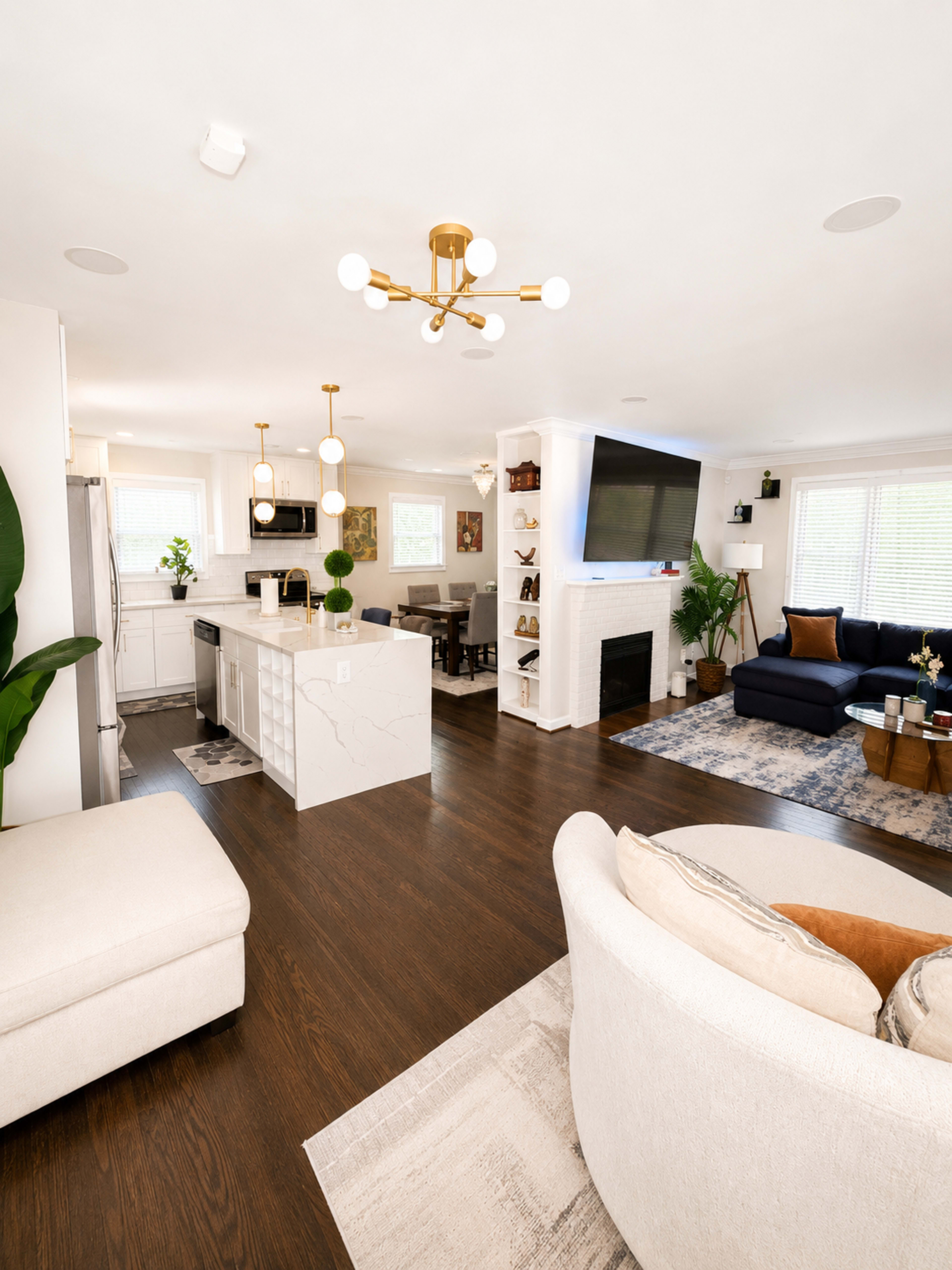 The image shows a modern open-concept living space featuring a kitchen with a large island, a dining area, and a cozy lounge with a fireplace and a television.