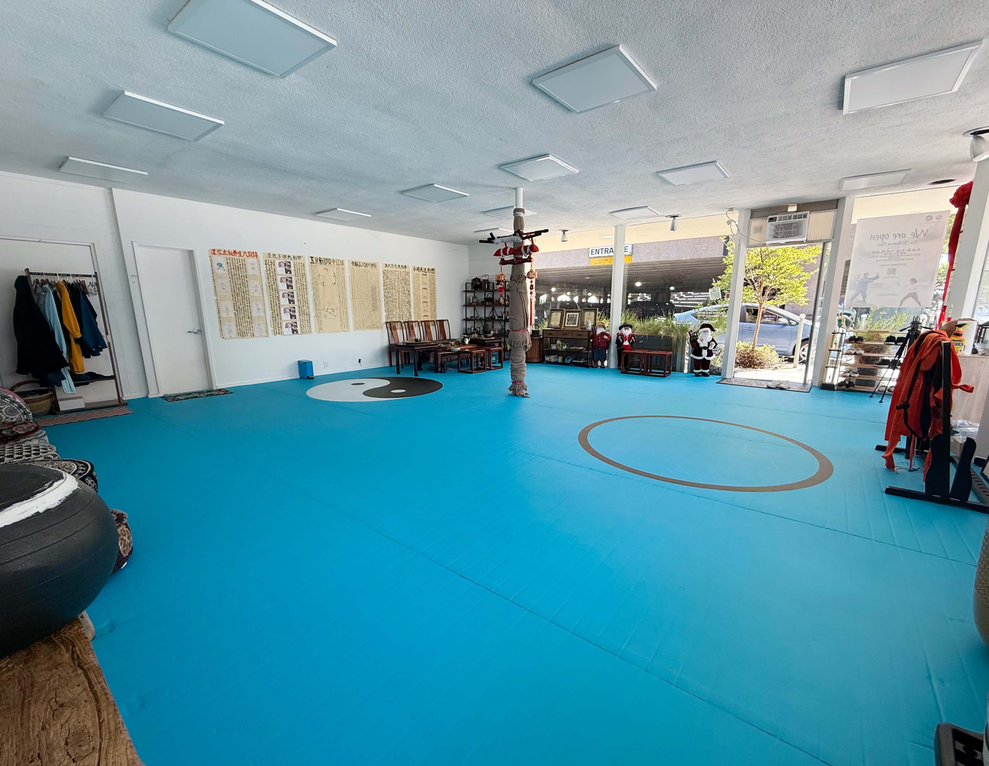 The image shows a spacious martial arts studio with blue flooring, wooden benches, and walls featuring various posters and charts.
