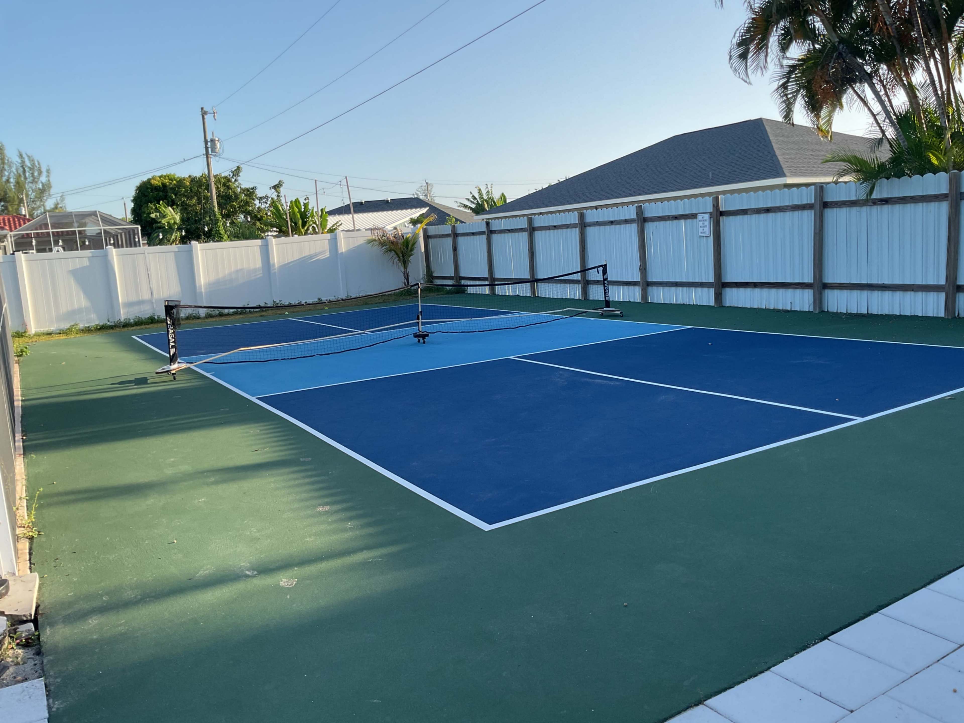 The image shows a tennis court with blue and green surfaces, surrounded by a white fence and palm trees.