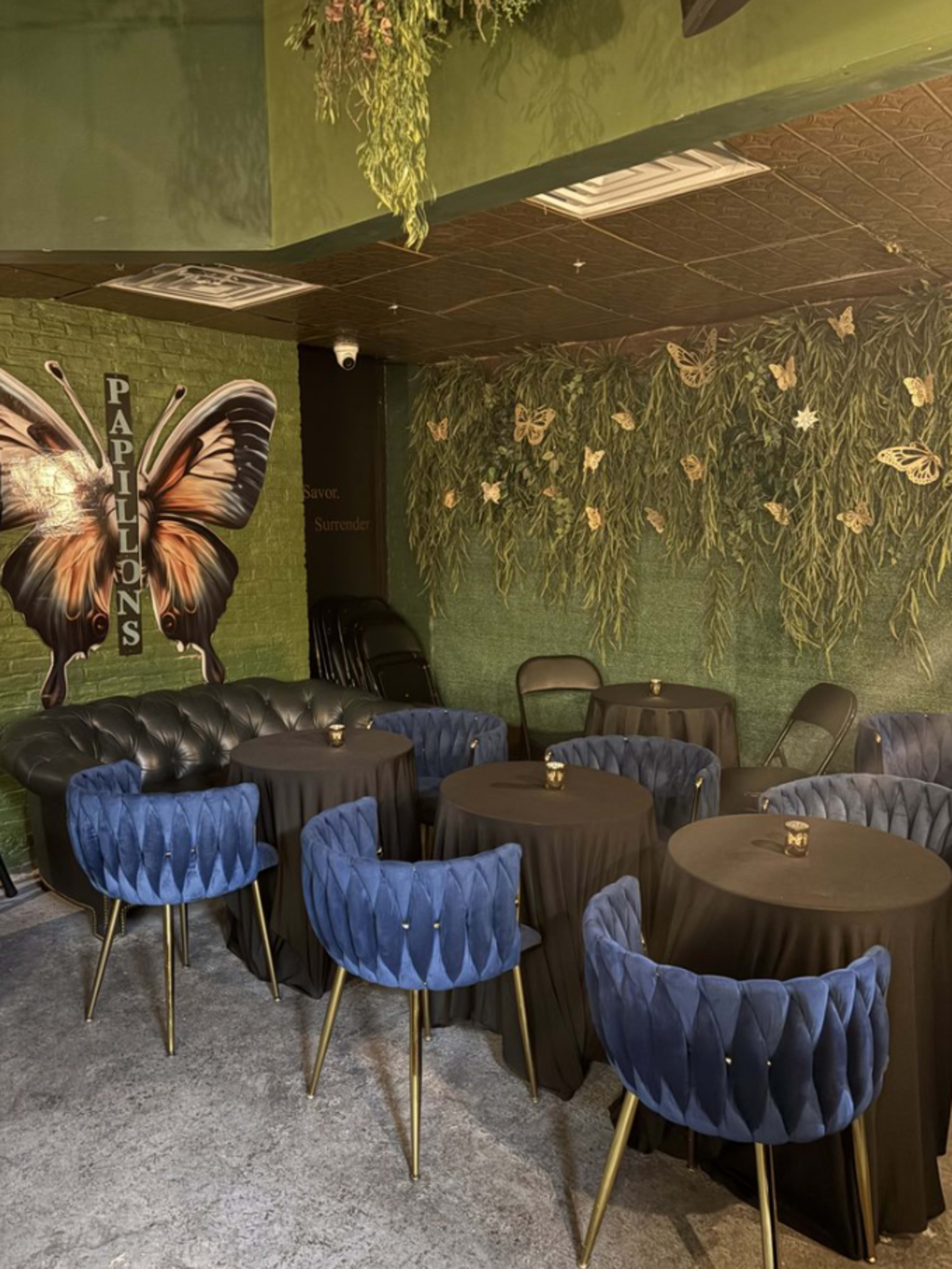 The image shows a cozy interior of a café or lounge, featuring a mural of a butterfly, black tables with dark cloths, and blue tufted chairs arranged around the space.