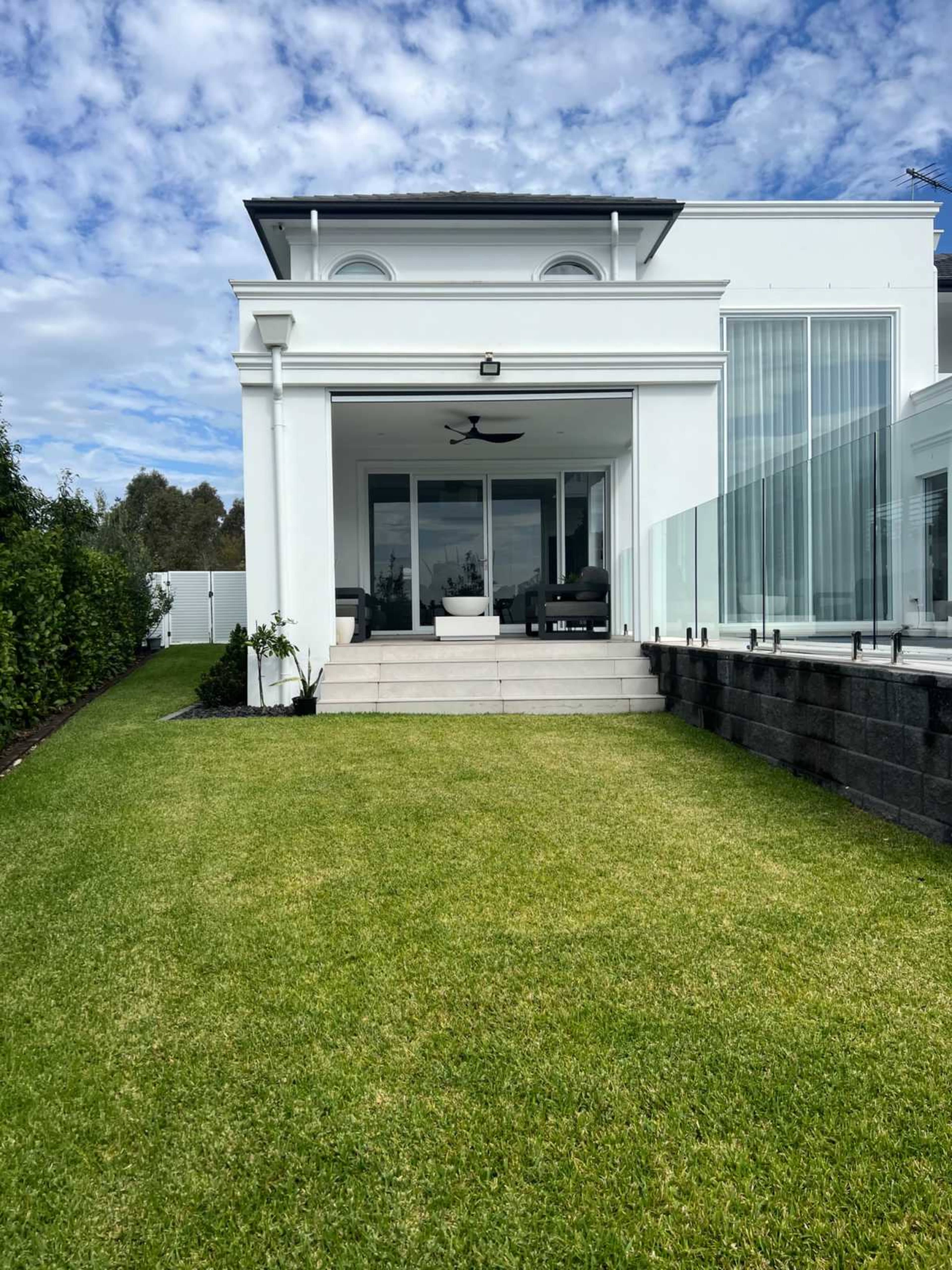 The image shows a modern white house with a spacious lawn and large glass doors leading to a patio area.