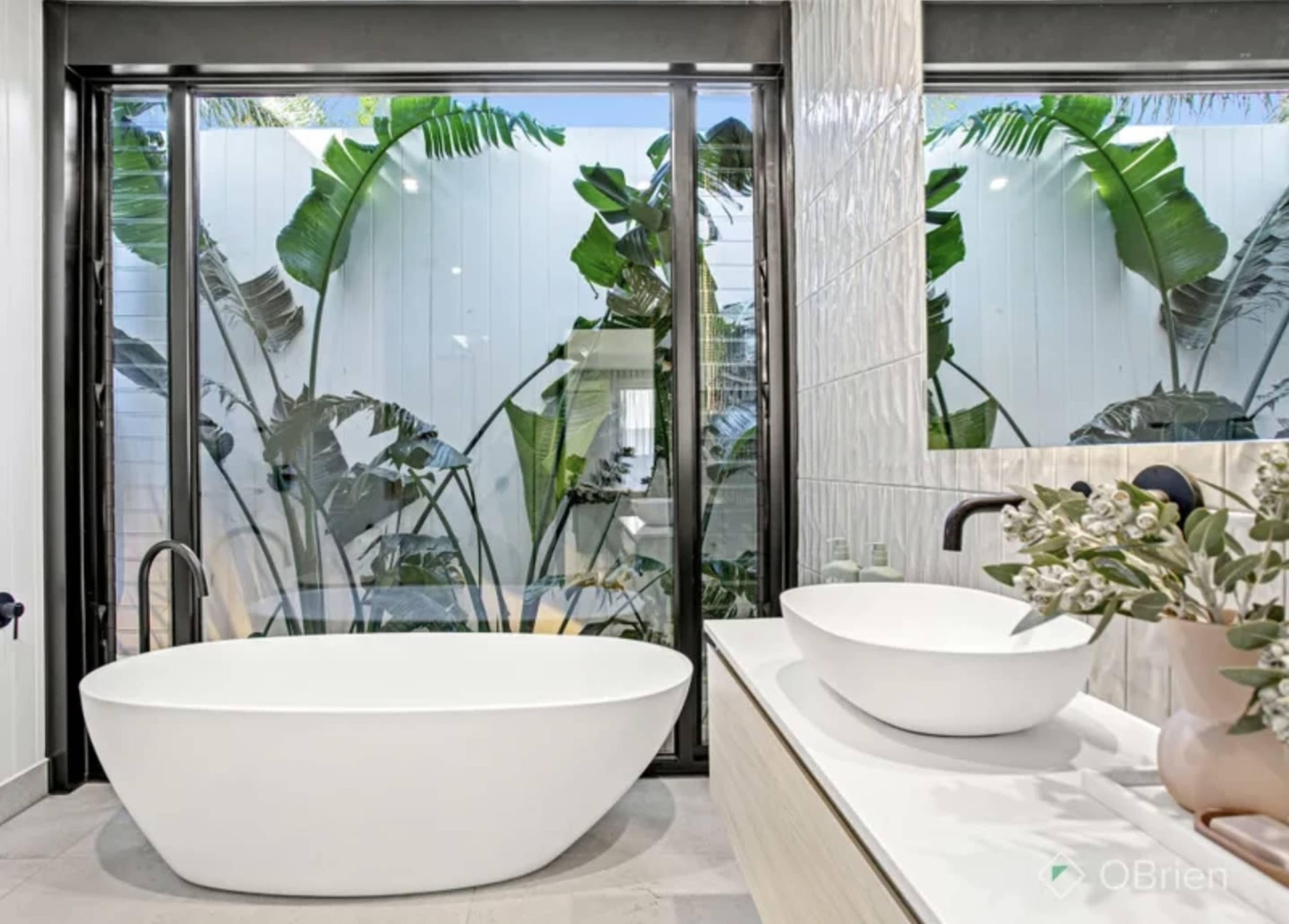 The image features a modern bathroom with a freestanding white bathtub, a double sink vanity, and tall glass windows showcasing greenery outside.