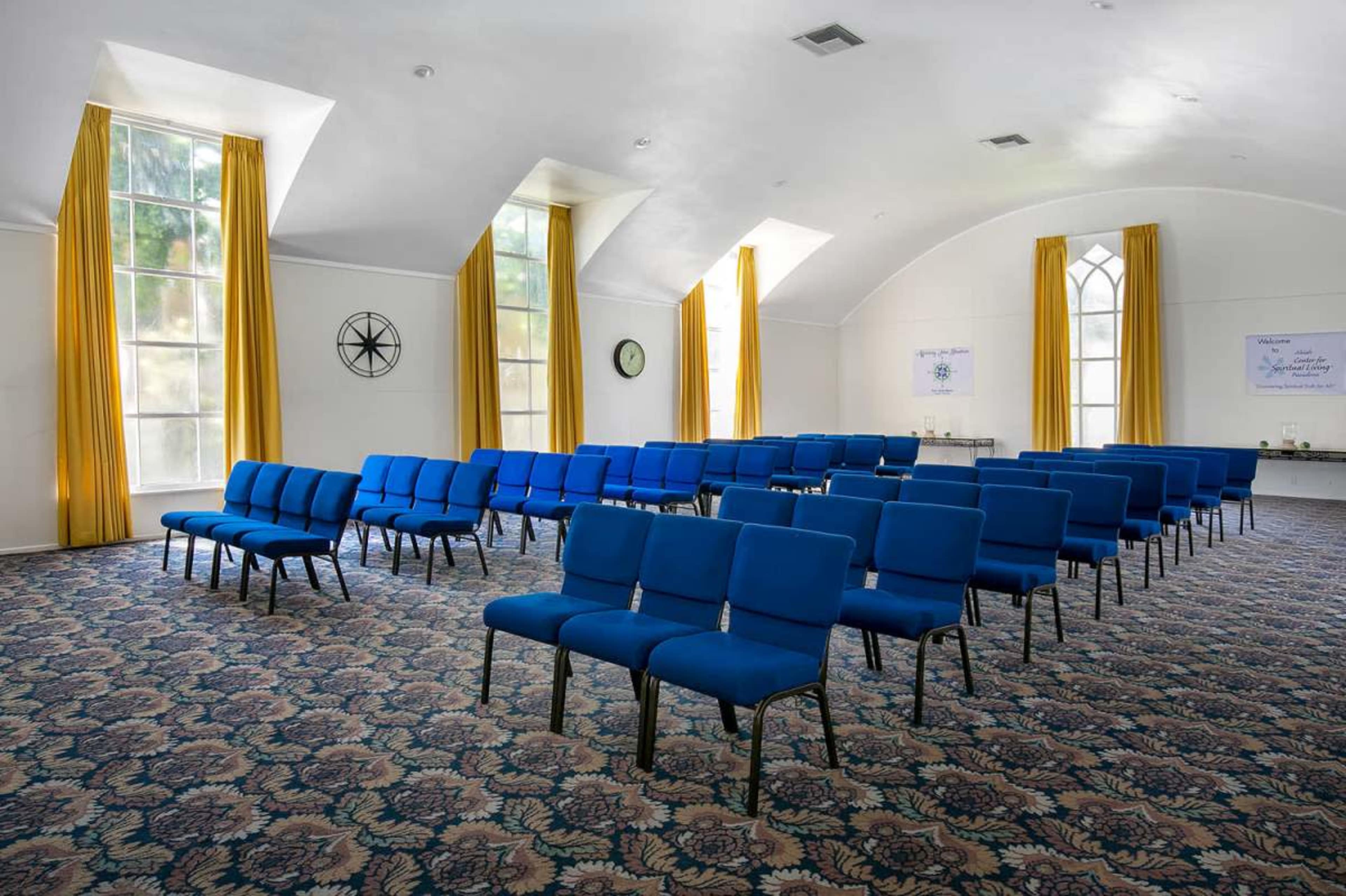 The image shows a large room with blue chairs arranged in rows on a patterned carpet, complemented by yellow curtains and clock on the wall.