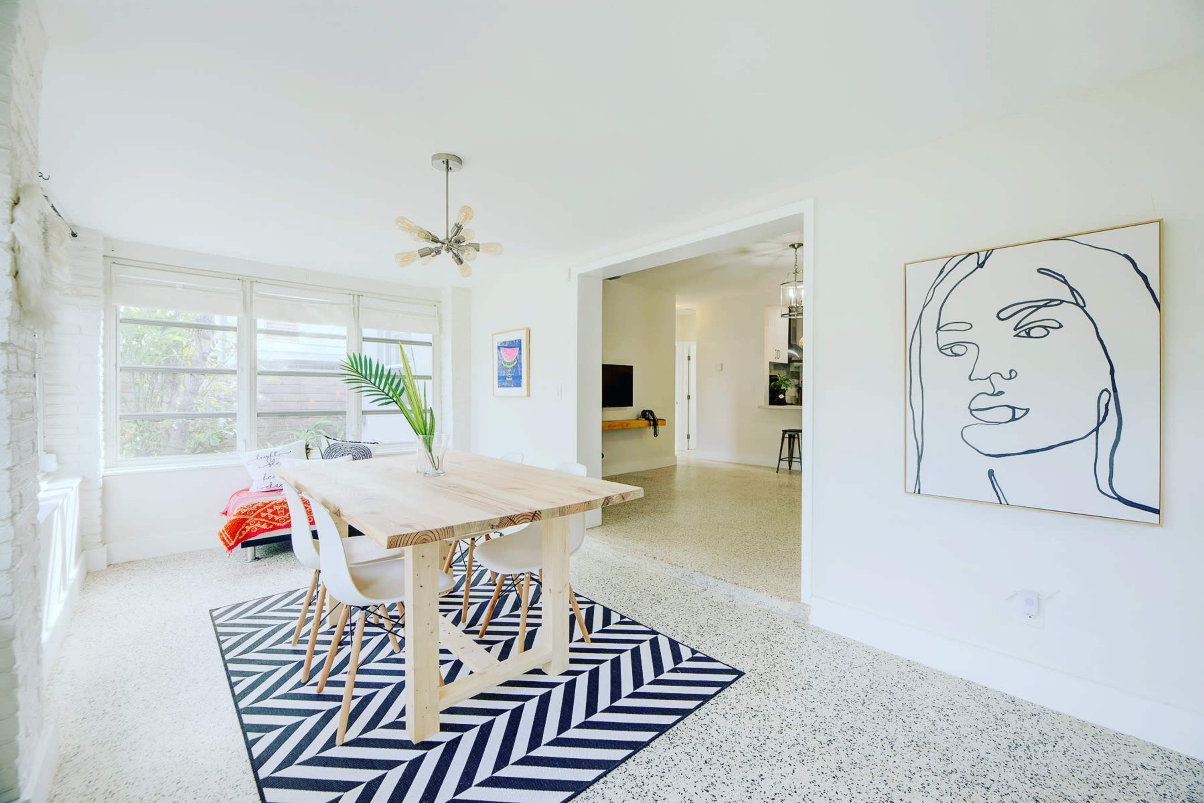 A dining area features a wooden table surrounded by white chairs, adorned with a modern abstract line drawing on the wall and a patterned rug beneath.