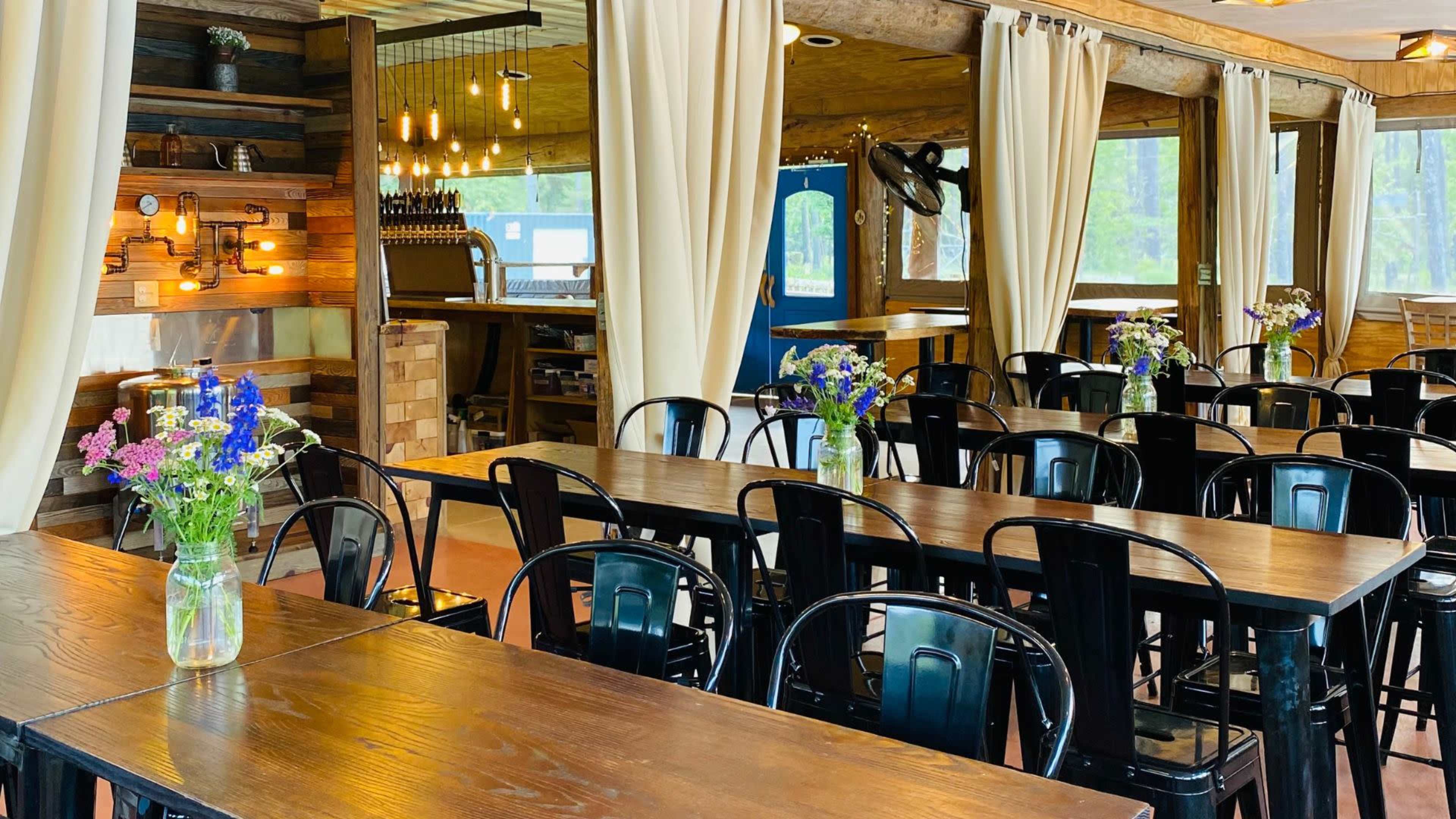 A dining area with wooden tables, black metal chairs, and flower vases on each table in a well-lit setting.