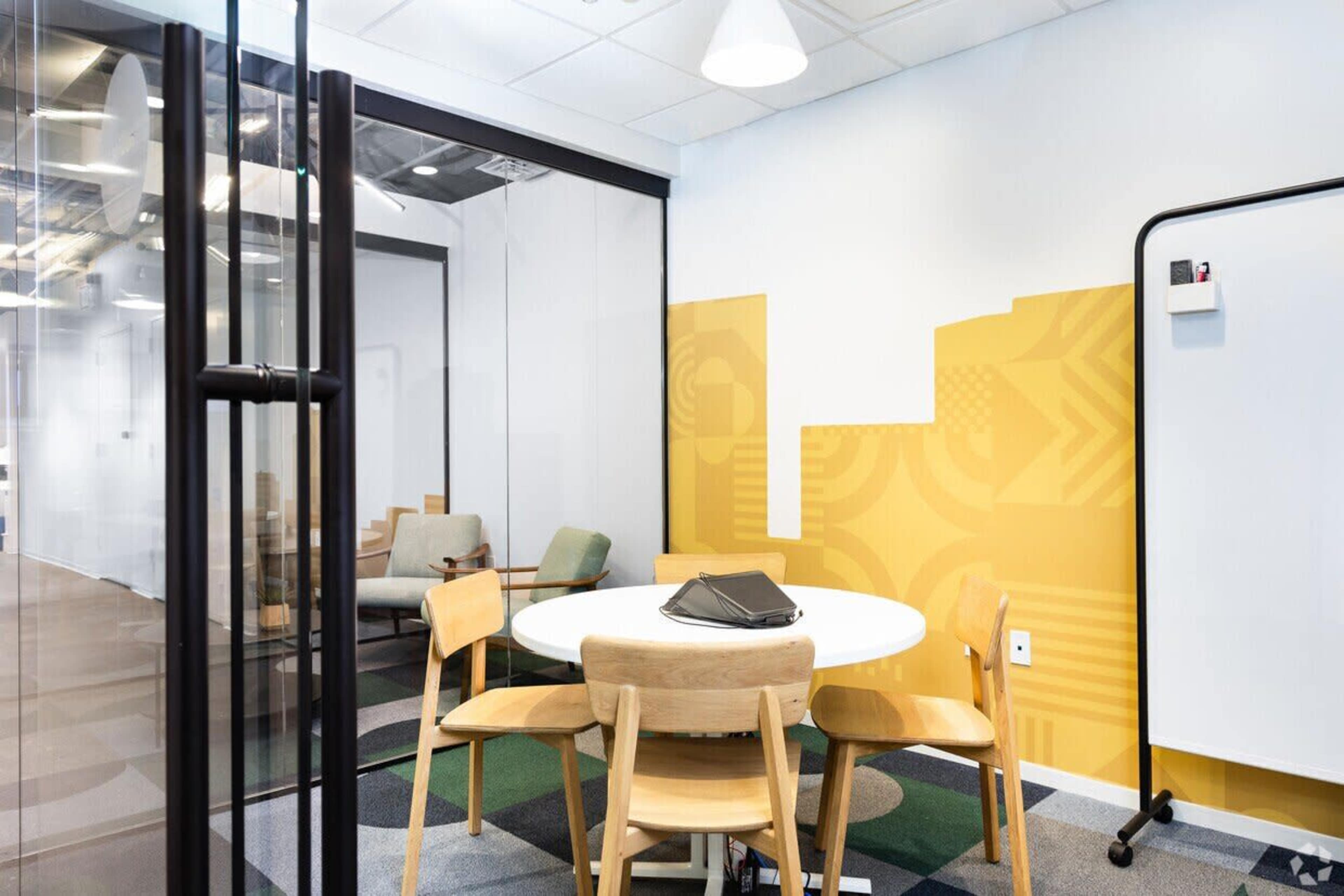 The image shows a modern conference room with a round table surrounded by four wooden chairs, a whiteboard, and a yellow accent wall.