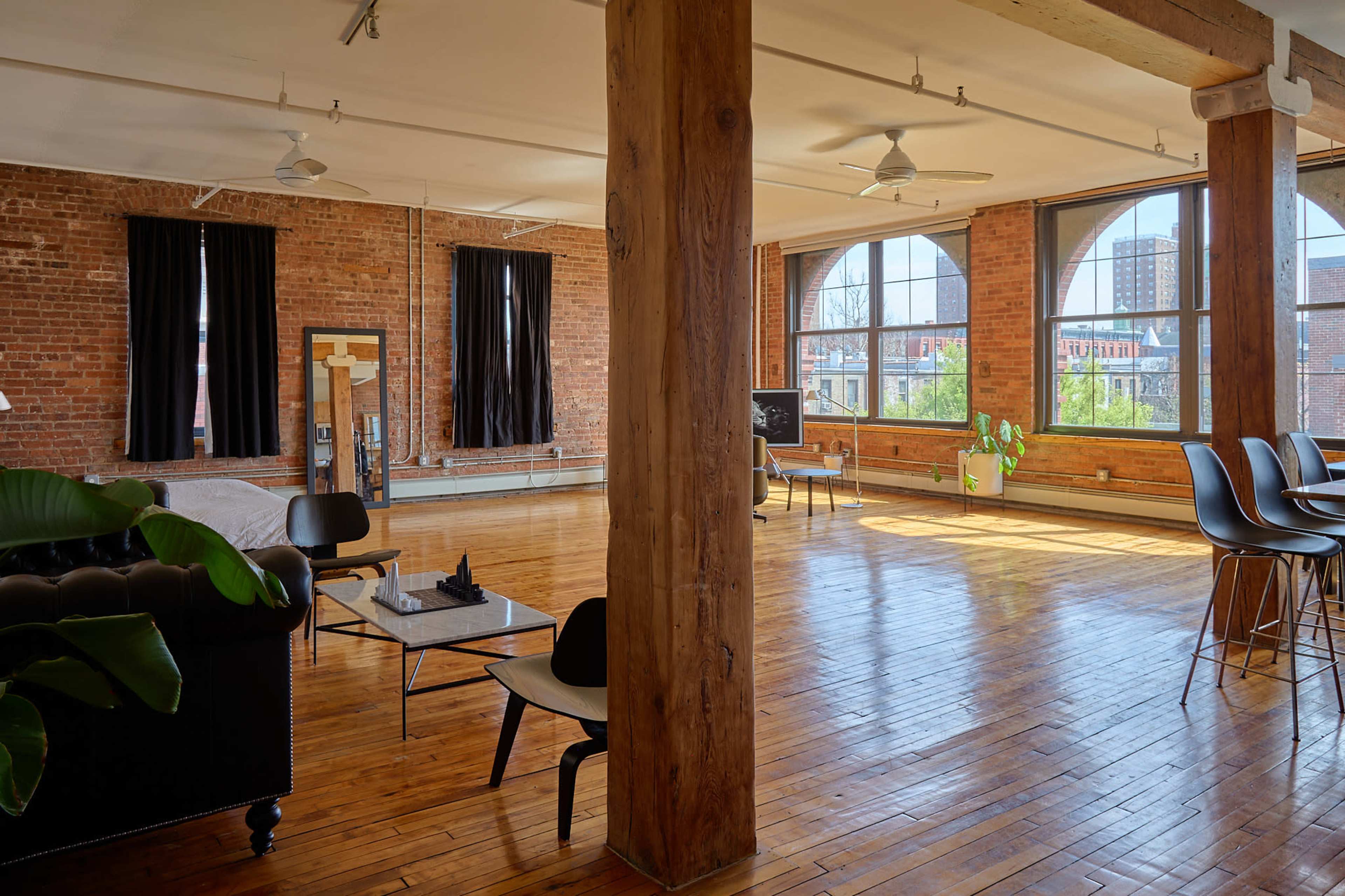 The image shows a spacious loft with exposed brick walls, large windows, wooden floors, and a mix of modern furniture.