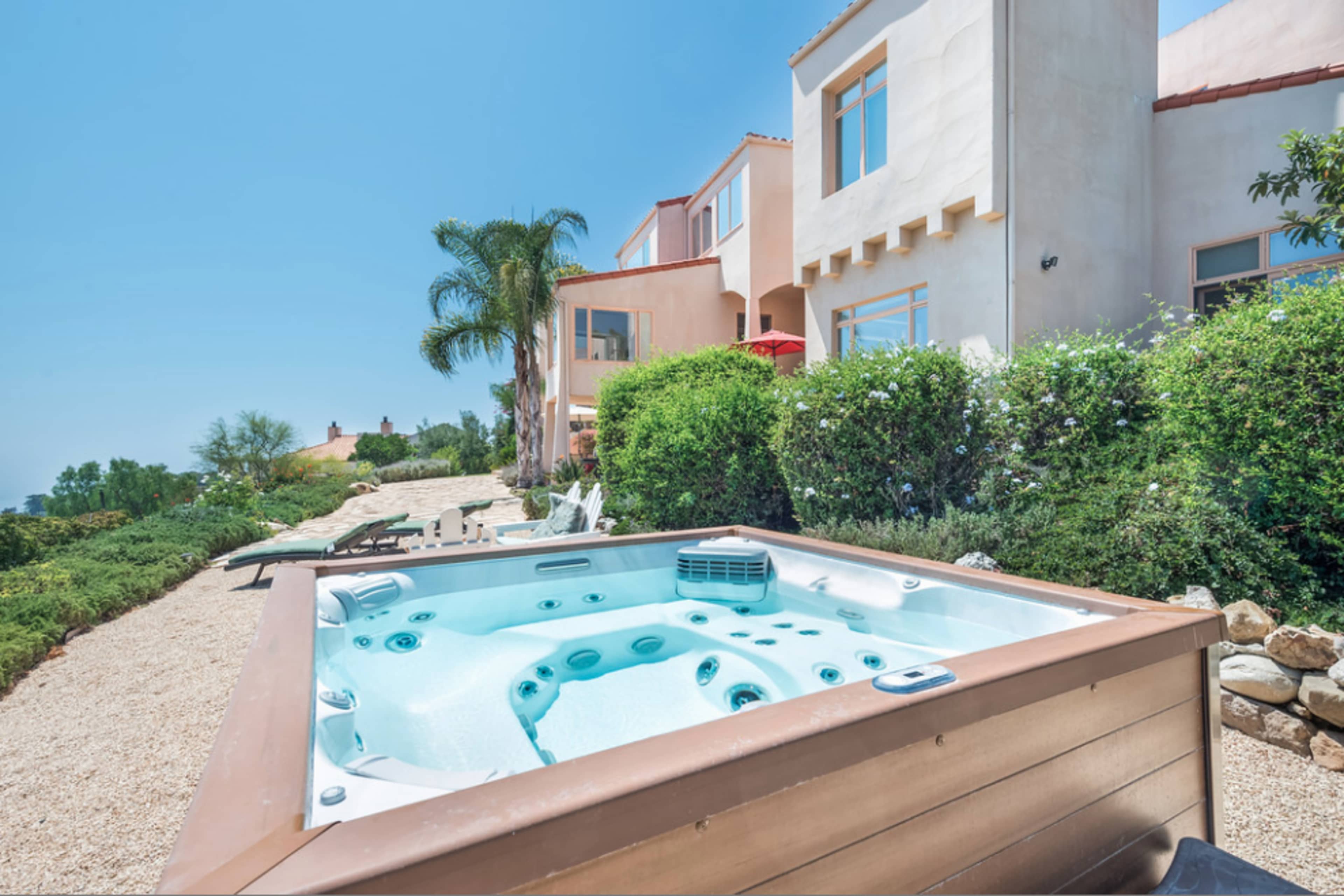 A hot tub sits in a landscaped yard with a view of a house and surrounding greenery.