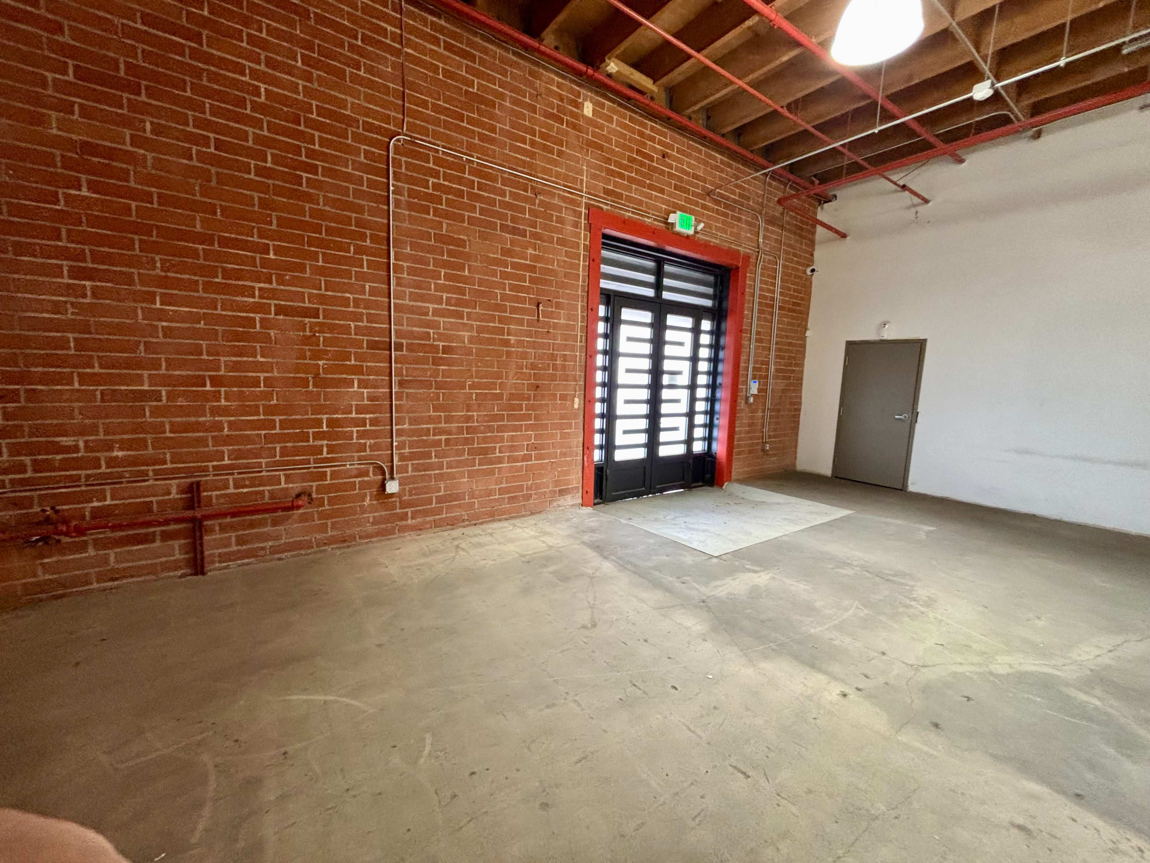The image shows an empty room with exposed brick walls, a double-door entrance, and a plain concrete floor.