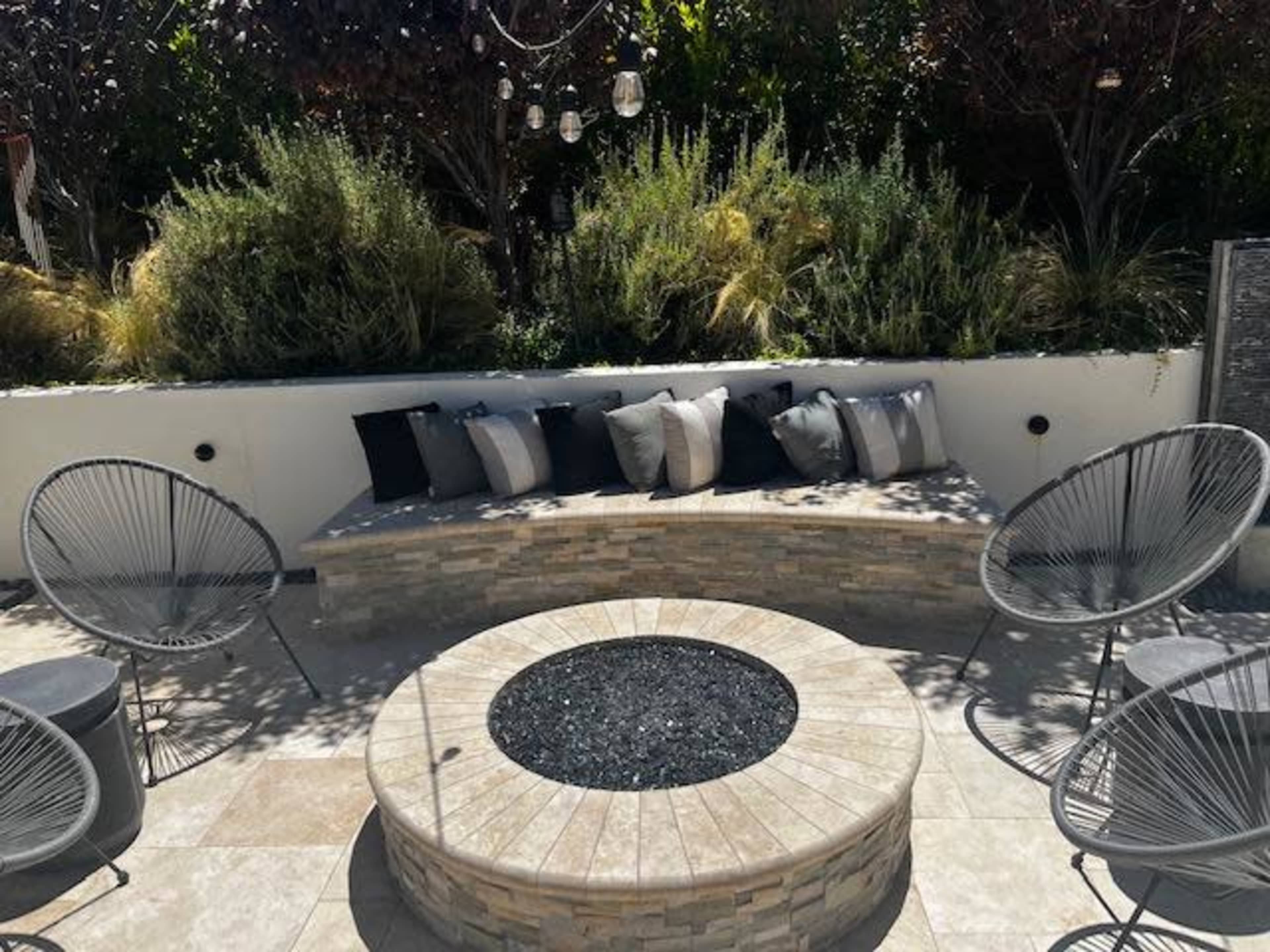 An outdoor seating area with a stone curved bench, decorative pillows, and a fire pit surrounded by modern chairs.