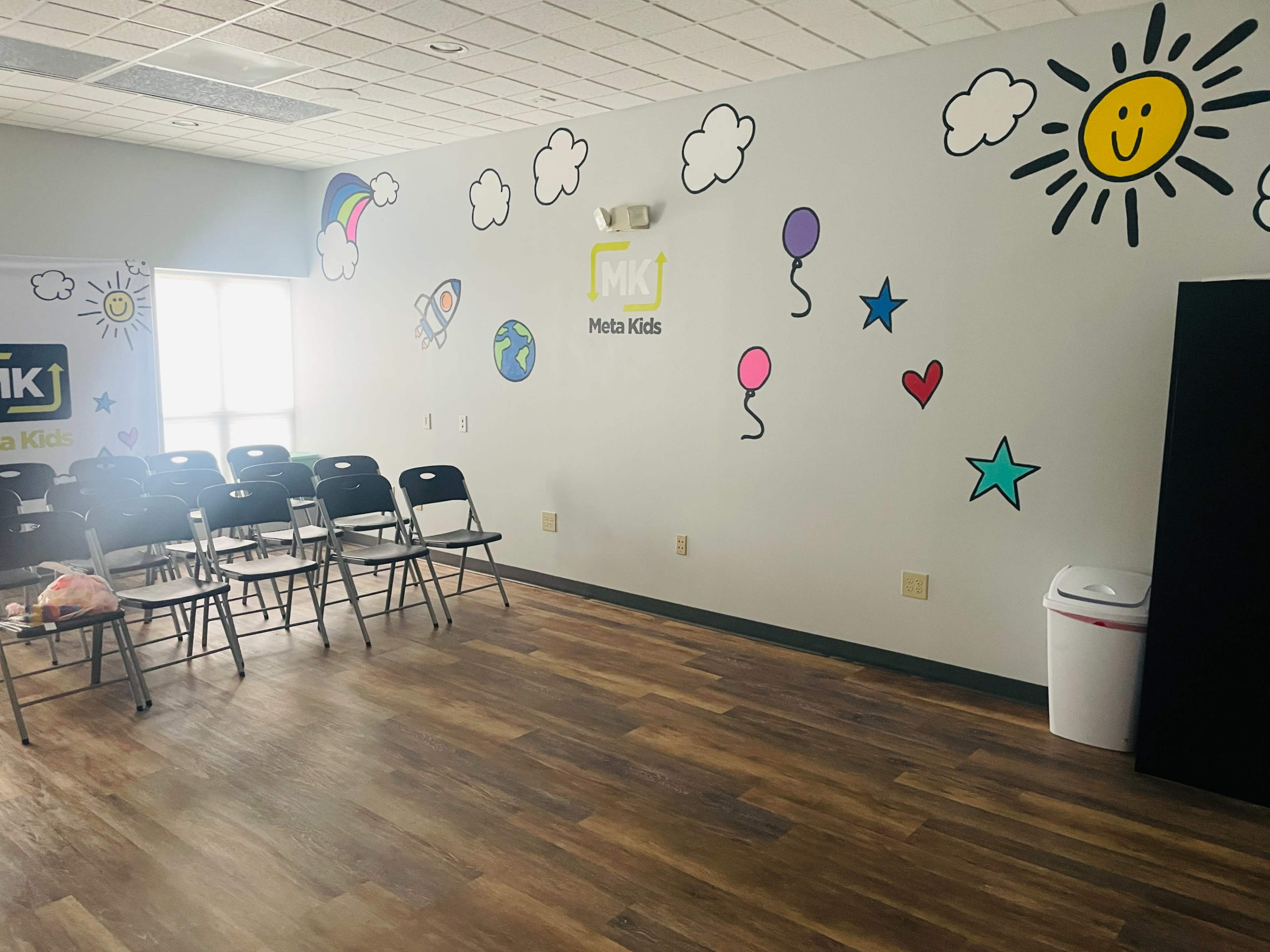 The room features several colorful wall decorations, including clouds, balloons, and a sun, with empty black chairs arranged in rows.