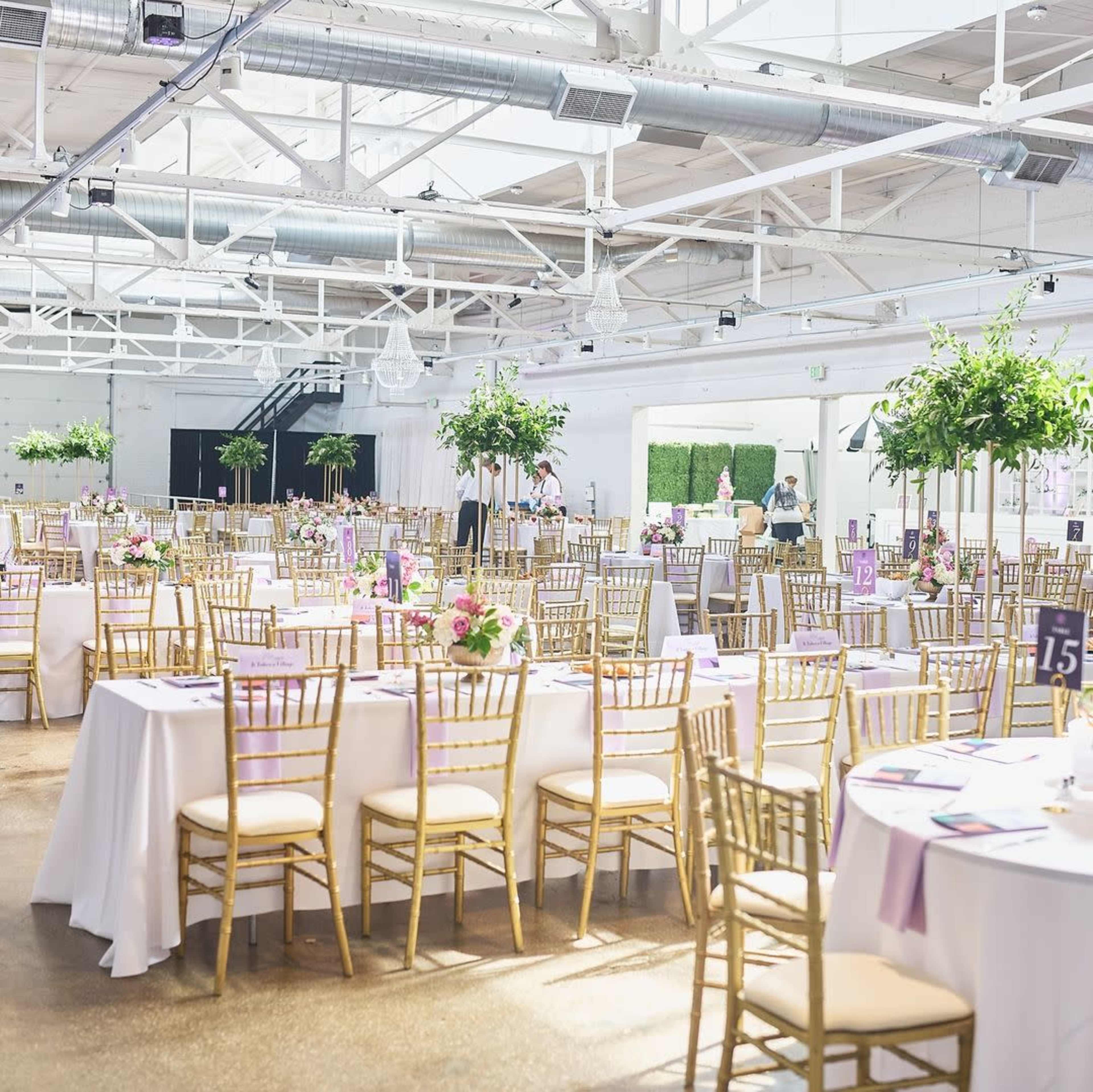 The image shows a spacious event venue arranged with tables covered in white linens, adorned with floral centerpieces and gold chiavari chairs.