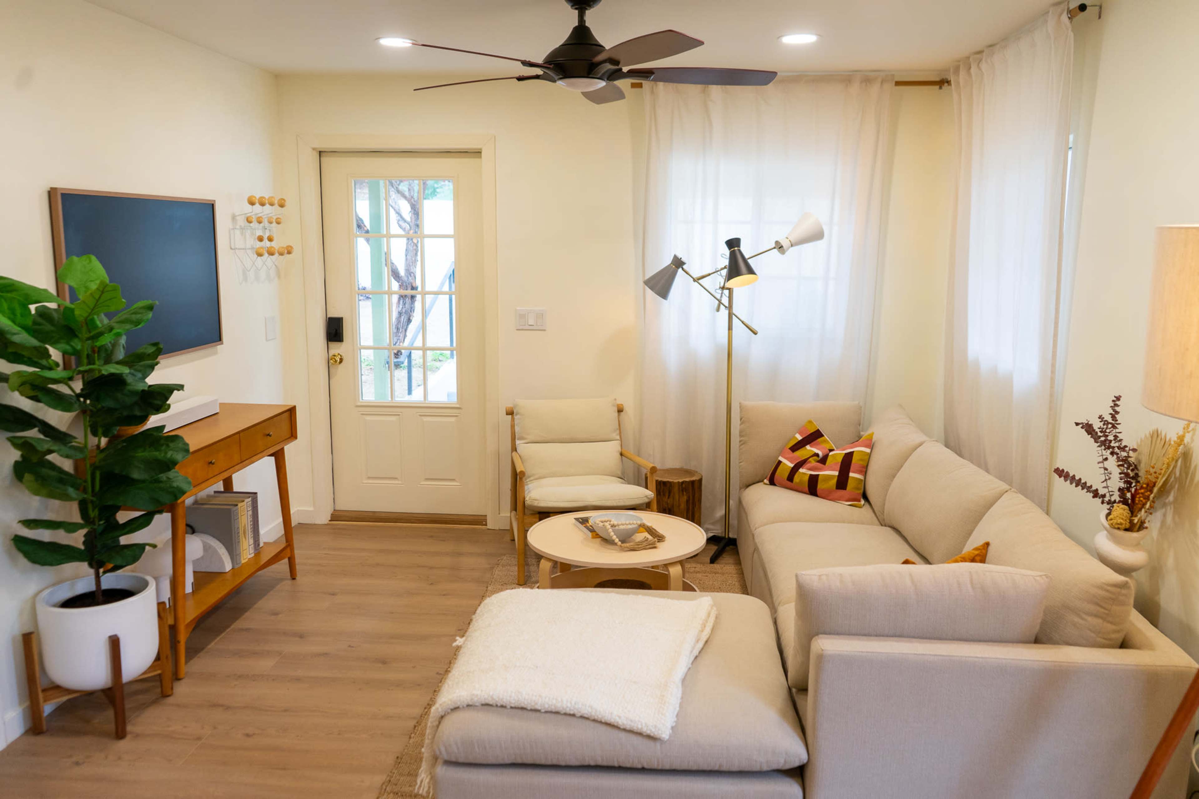 The image shows a bright living room with a sofa, two chairs, a coffee table, and a green plant near a door with a window.
