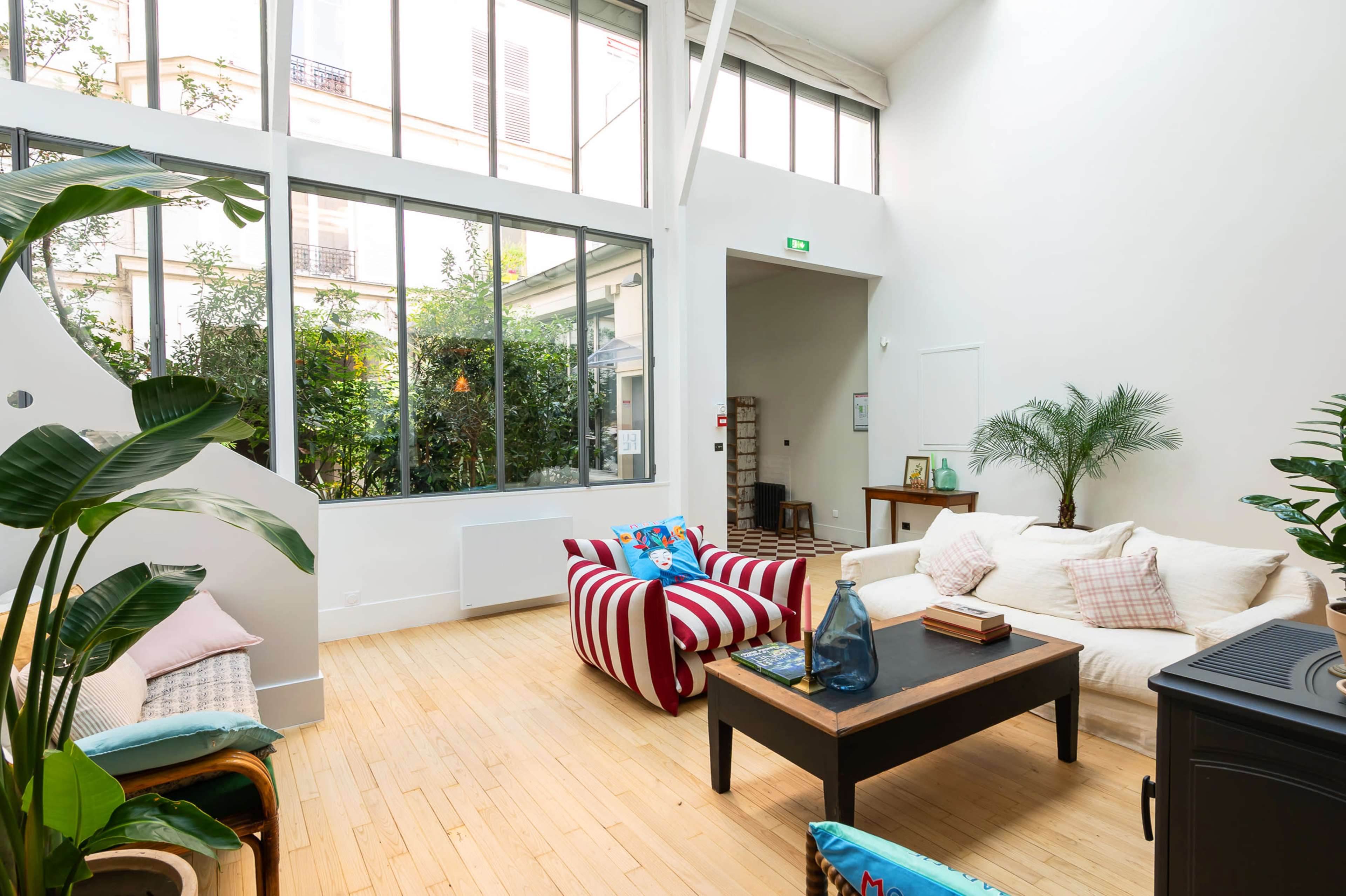 The image shows a bright, airy living room with large windows, a striped armchair, a coffee table, and various indoor plants.