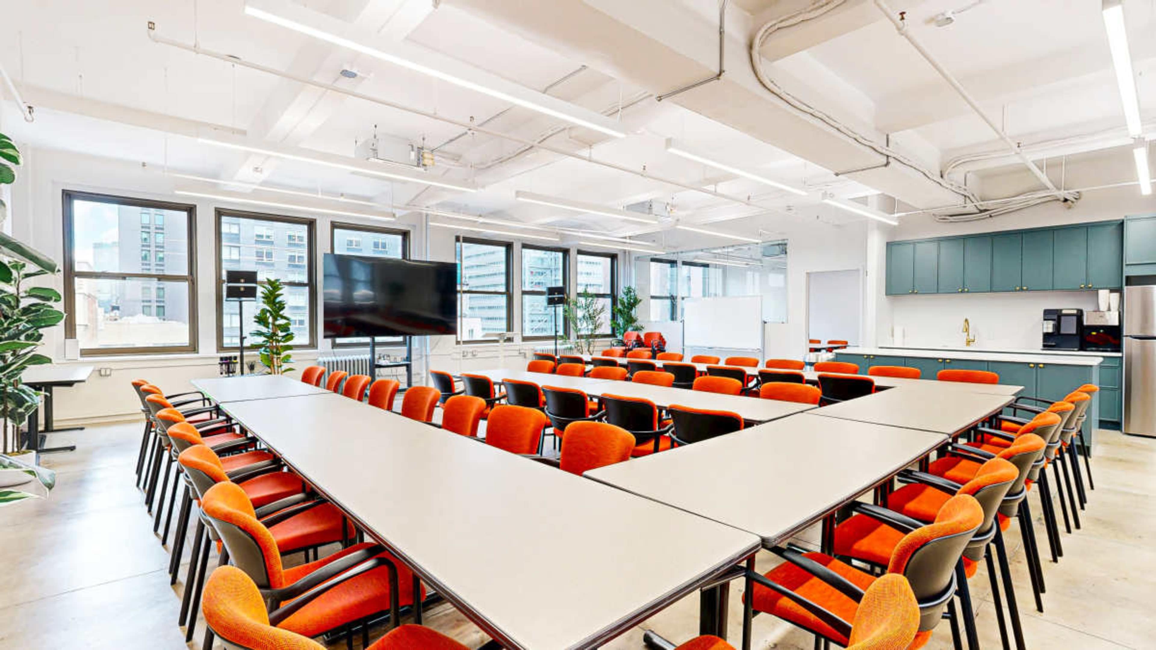 A spacious conference room features multiple long tables arranged in a V-shape, surrounded by orange chairs, with large windows allowing natural light and a kitchenette in the background.