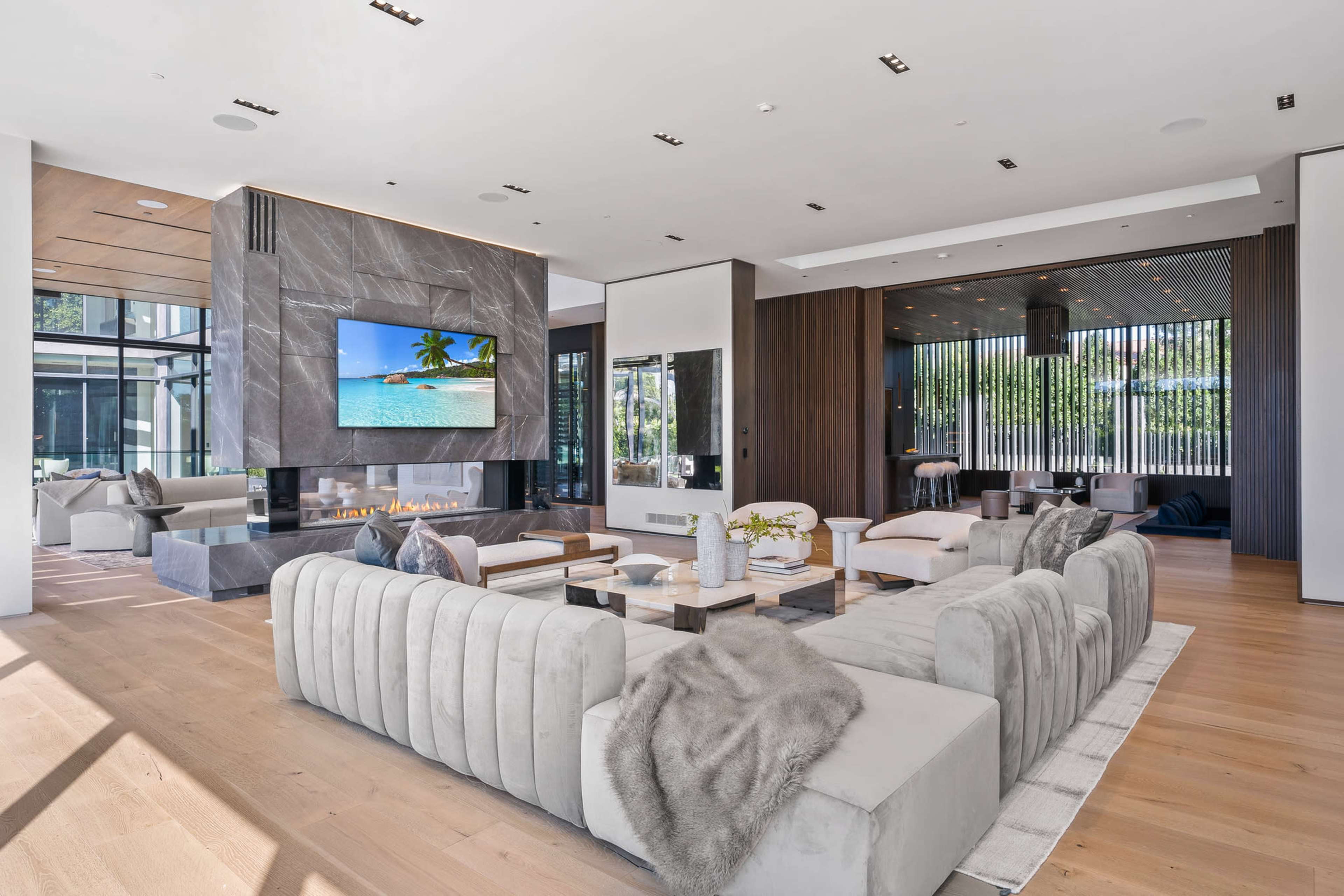 The image showcases a modern living room with a large sectional sofa, a sleek fireplace, and a wall-mounted television, surrounded by floor-to-ceiling windows.