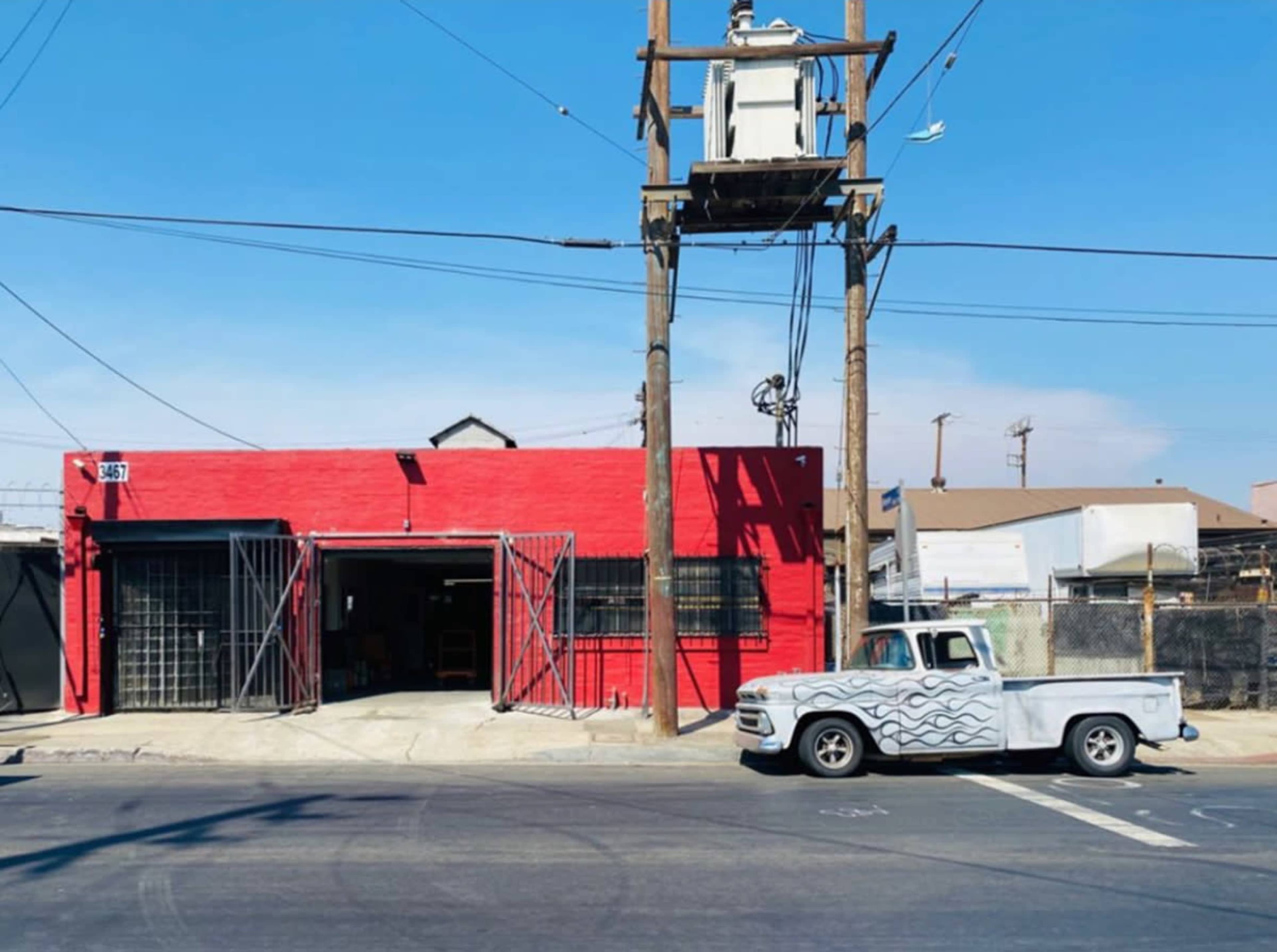 A red, industrial building with a large open doorway sits next to a wooden utility pole and a parked white truck with a wave design.