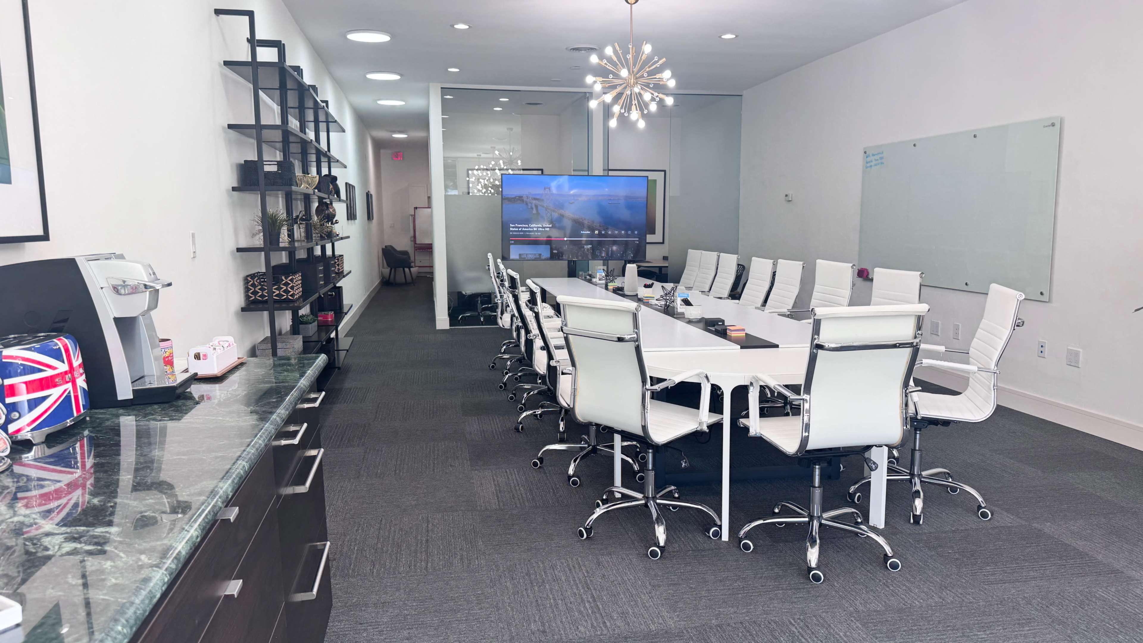The image shows a modern conference room with a long white table, ergonomic chairs, a large television screen, and a kitchen area along one side.
