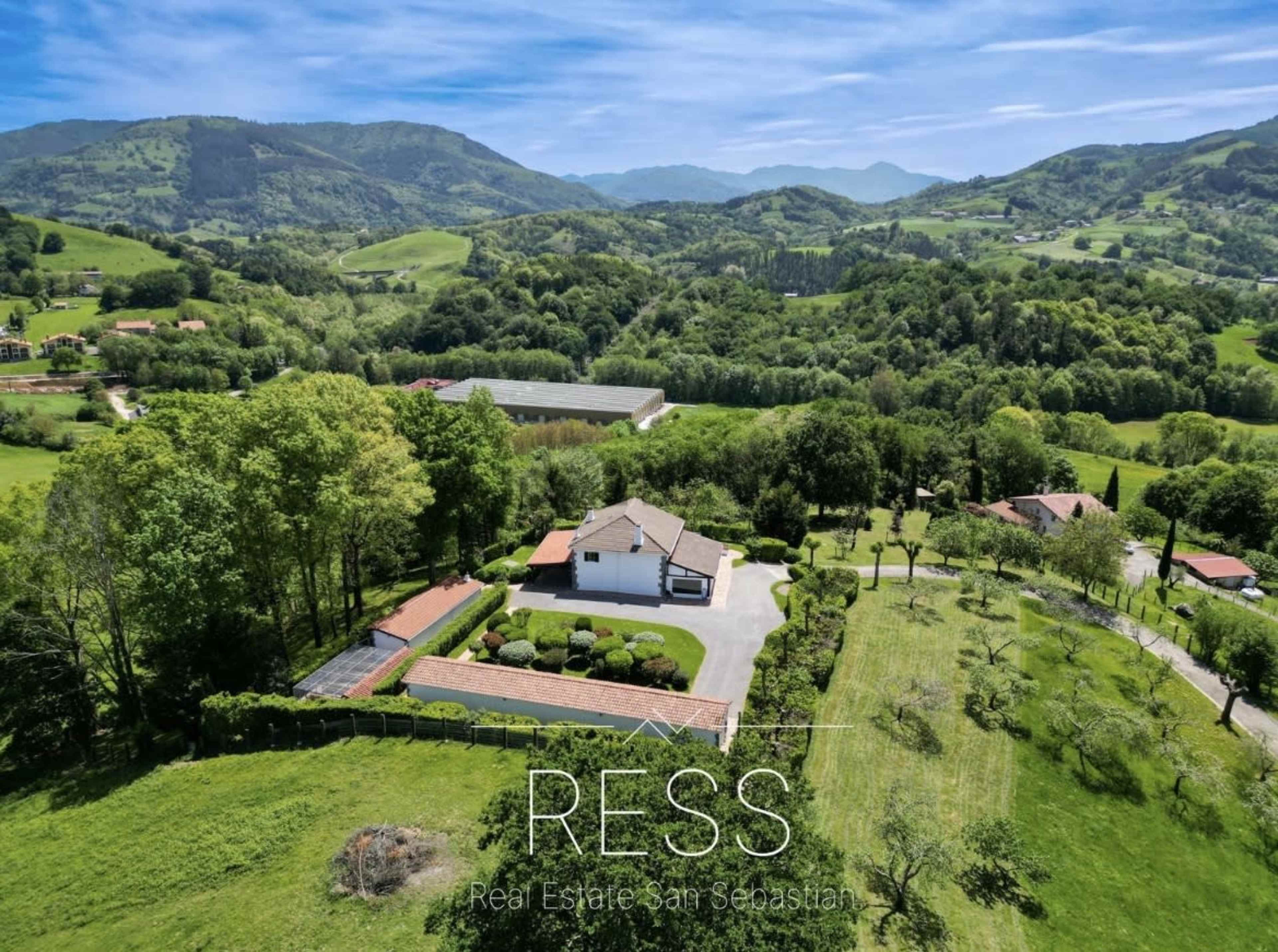 The image shows a picturesque landscape featuring a white house surrounded by greenery and rolling hills under a clear blue sky.