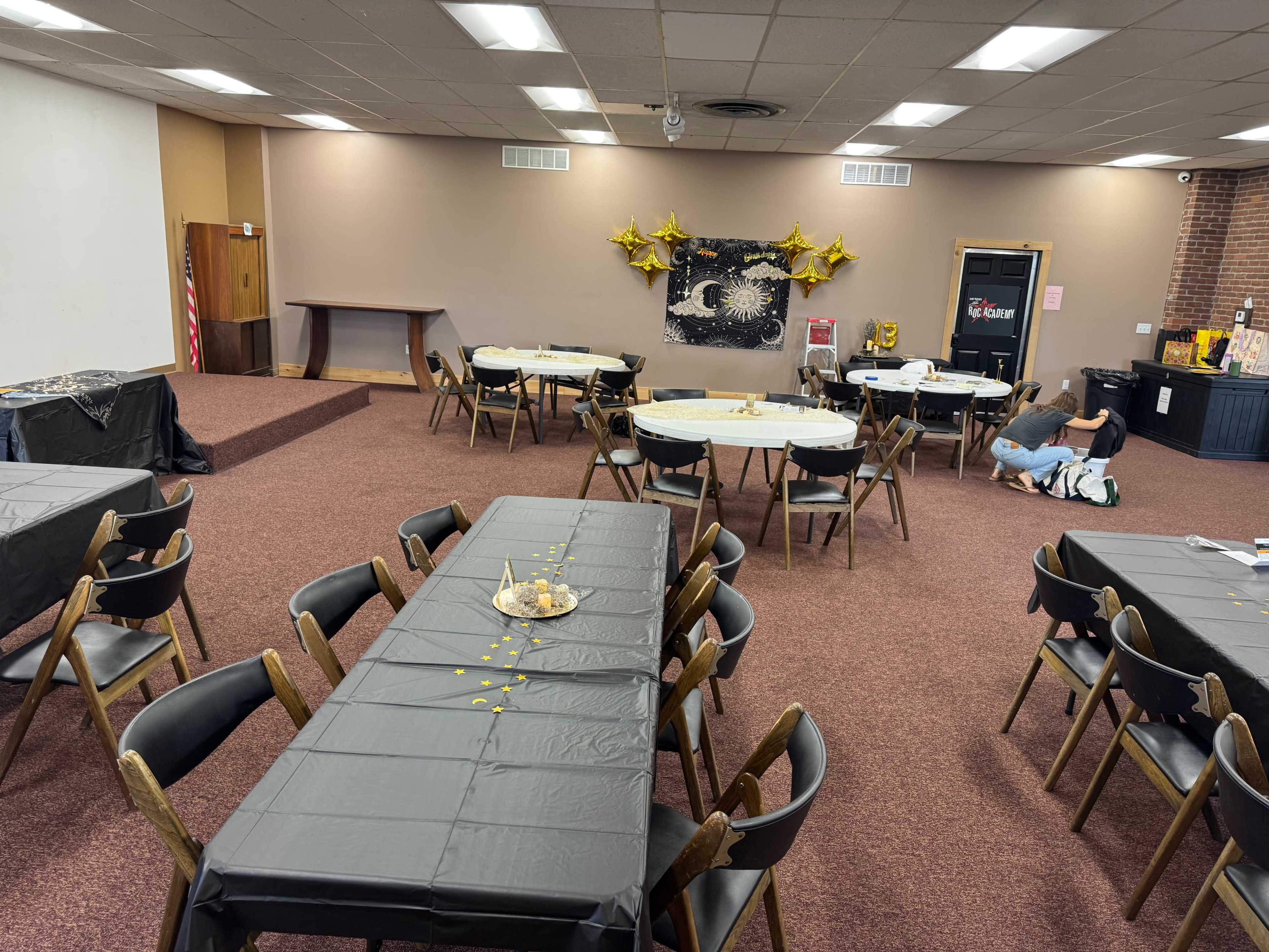 A large room is set up with multiple black tablecloth-covered tables arranged for an event, featuring gold star decorations on the wall.