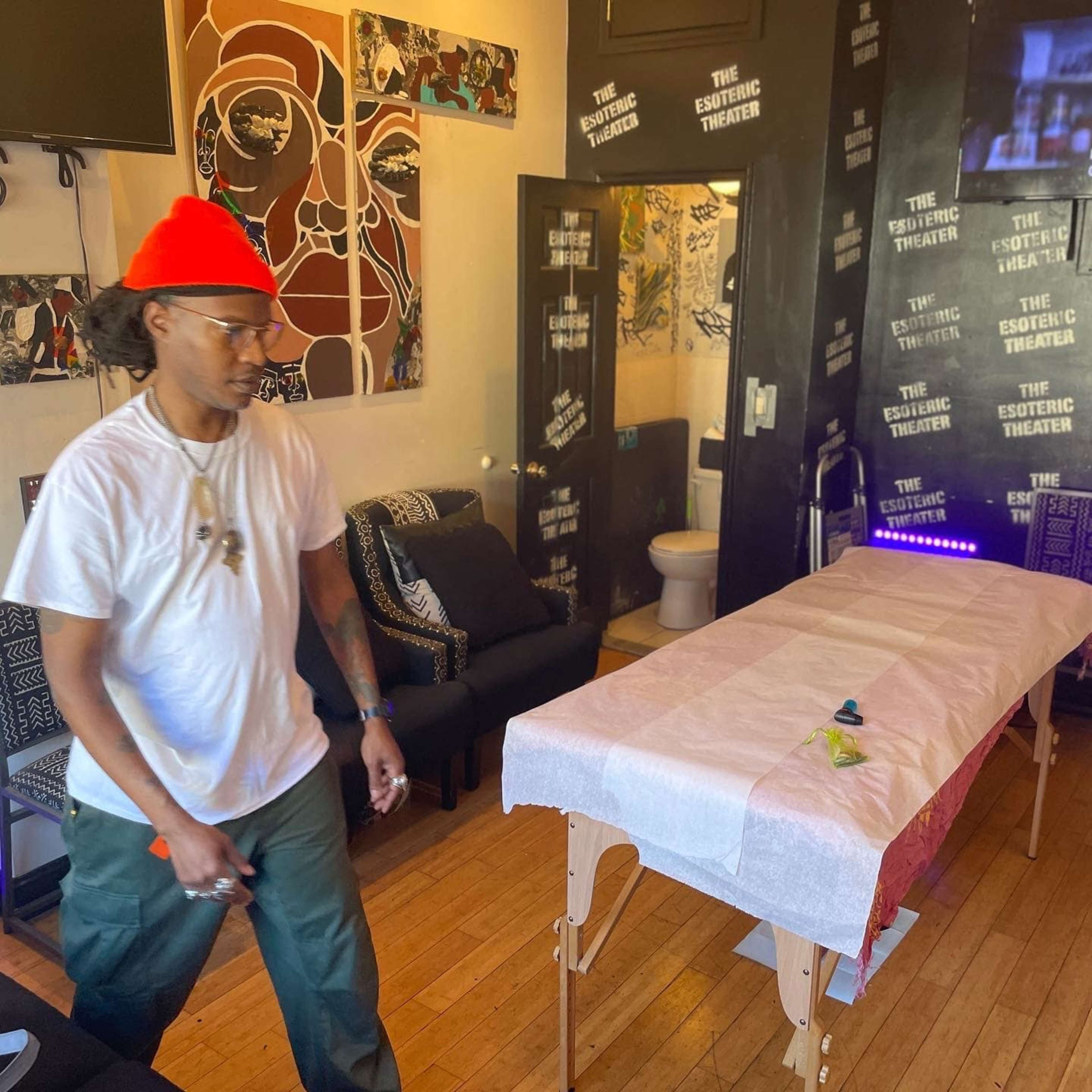 A person wearing a white shirt and an orange beanie walks past a massage table in a room decorated with artwork and black walls featuring text.