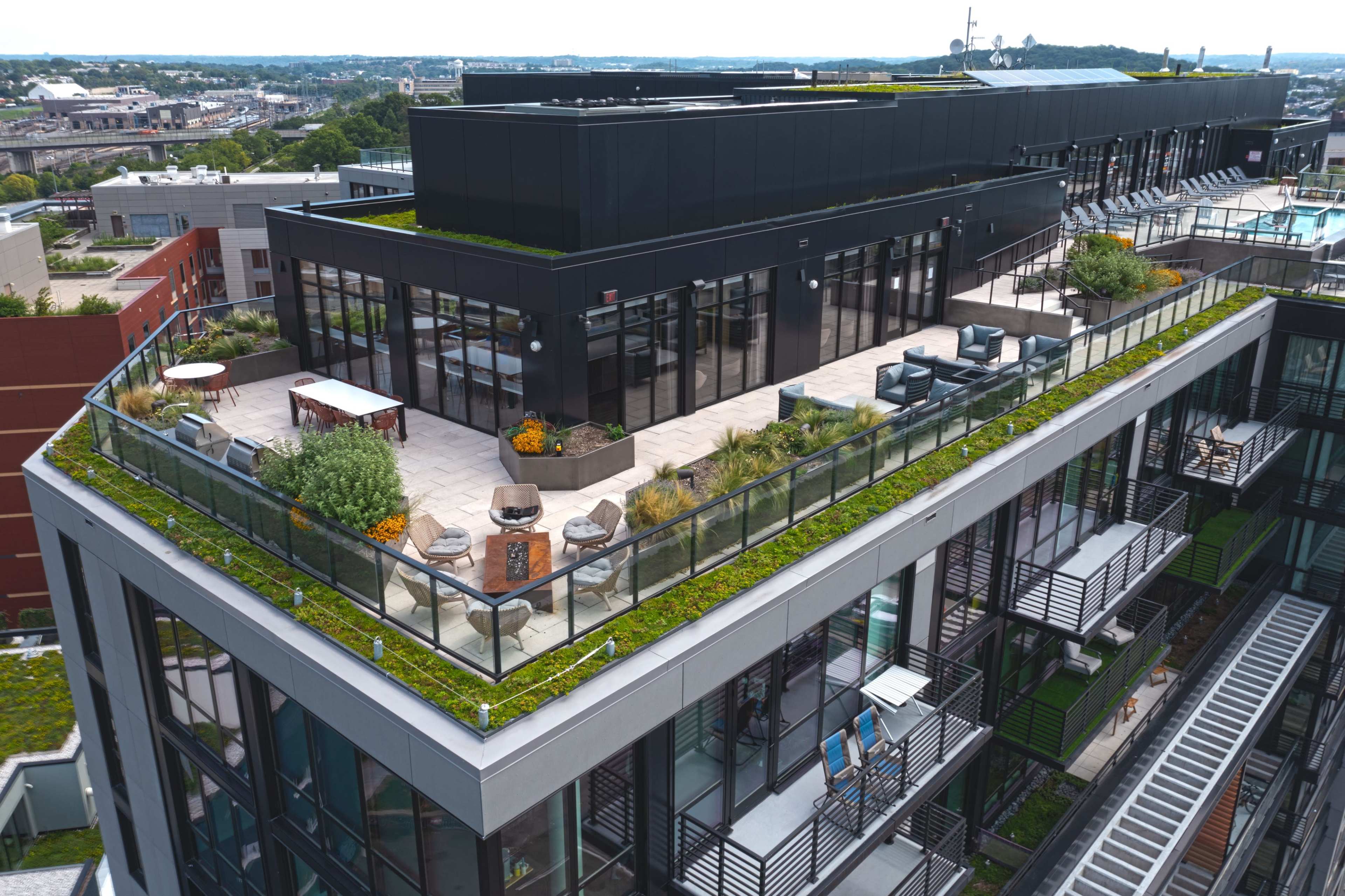 The image shows a rooftop terrace featuring greenery, seating areas, and urban views from a modern building.