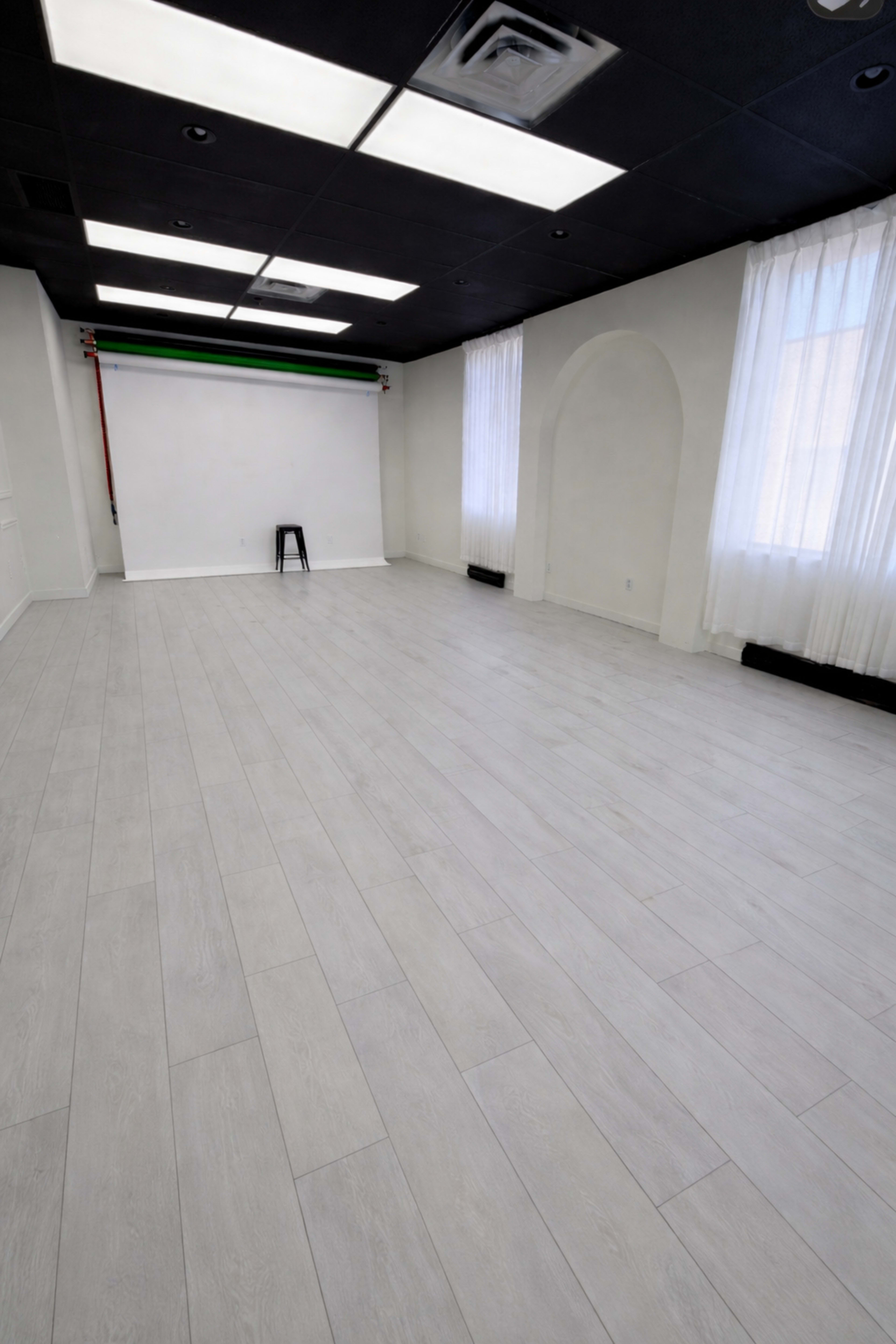 The image shows an empty, well-lit studio space with light-colored wooden flooring and large windows draped with sheer white curtains.