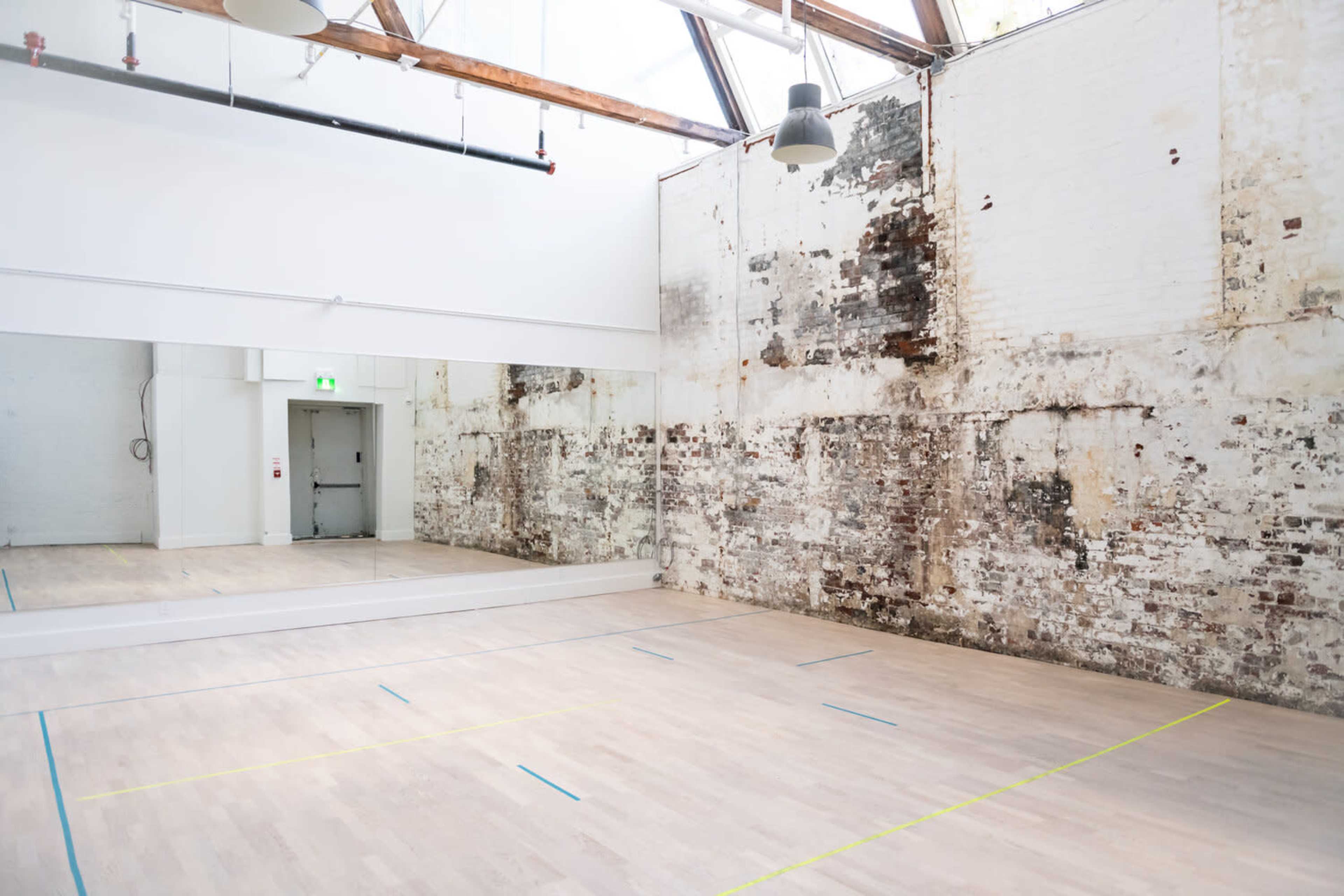 A spacious dance studio with wooden floors, a large mirrored wall, and unfinished brick walls, illuminated by overhead lighting and natural light from a skylight.