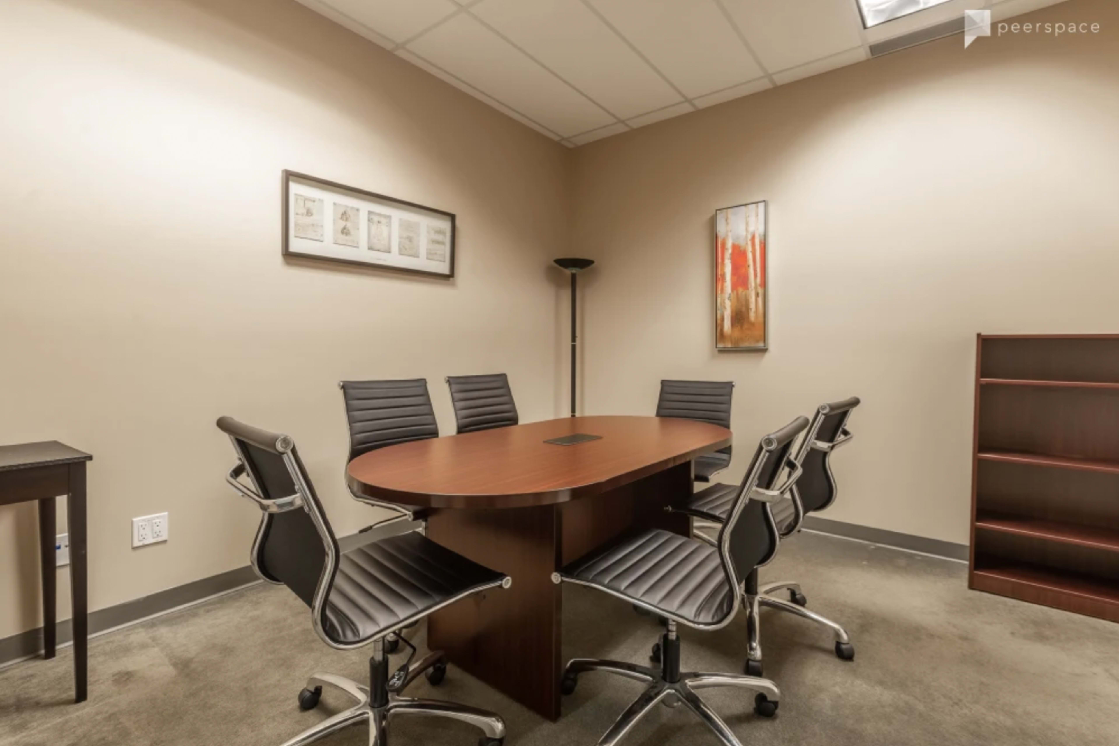 A small conference room featuring a round wooden table surrounded by six black office chairs, with a framed artwork on the wall and a tall lamp in the corner.