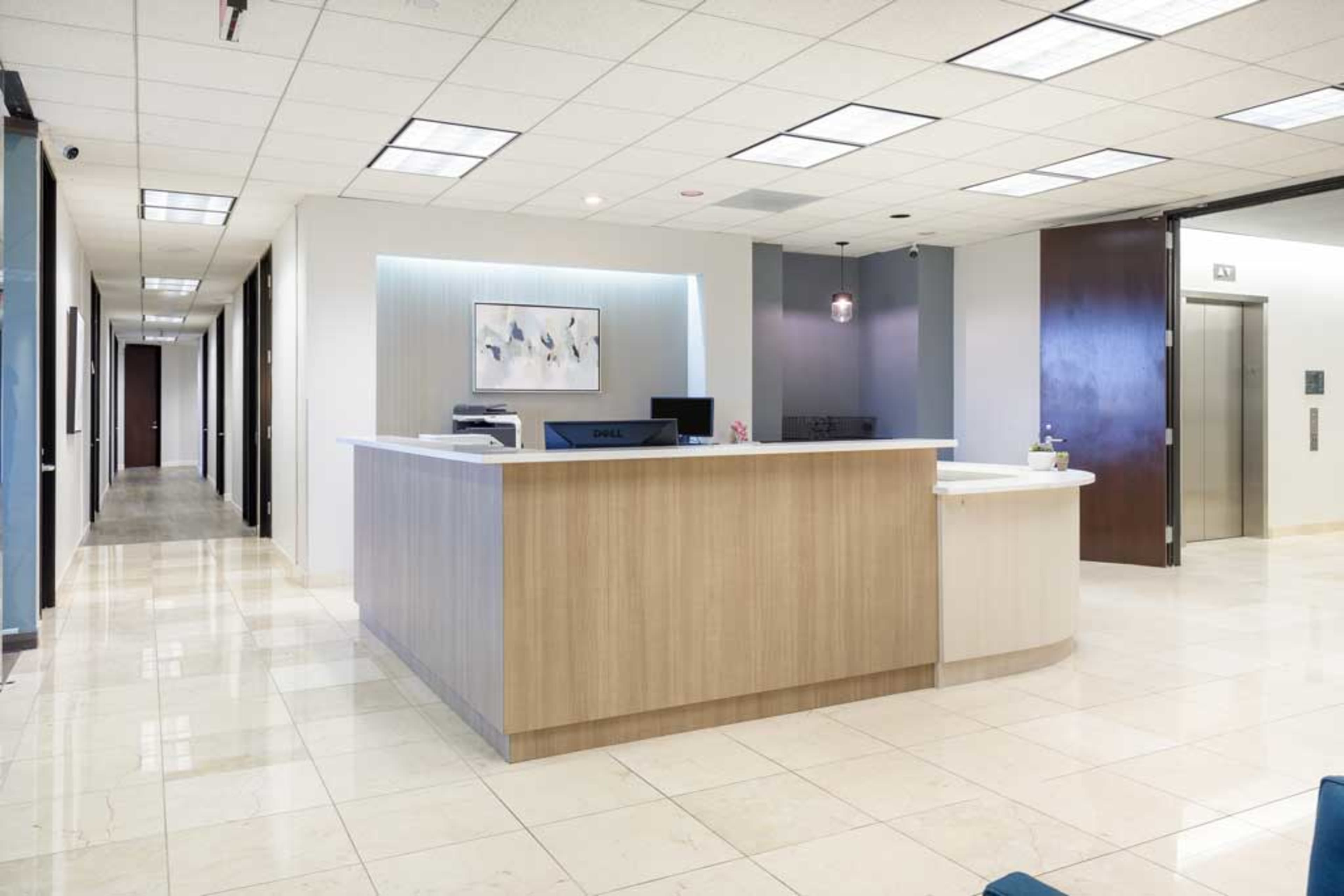 A modern reception area with a wooden front desk, an elevator to the right, and a well-lit hallway leading to office doors.