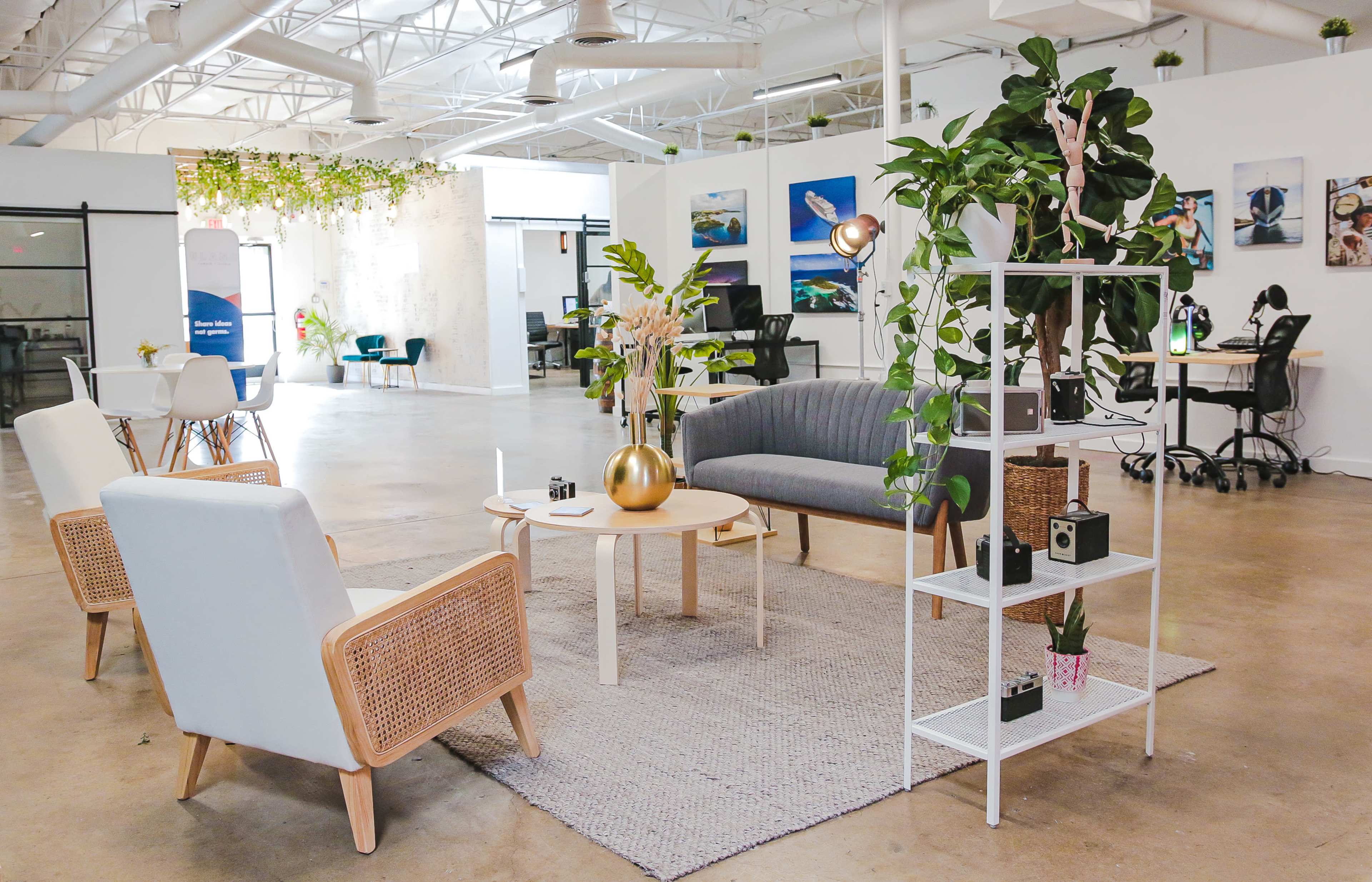 The image shows a modern workspace with a seating area featuring two armchairs, a coffee table, and a shelf with plants and decorative items, surrounded by workstations.