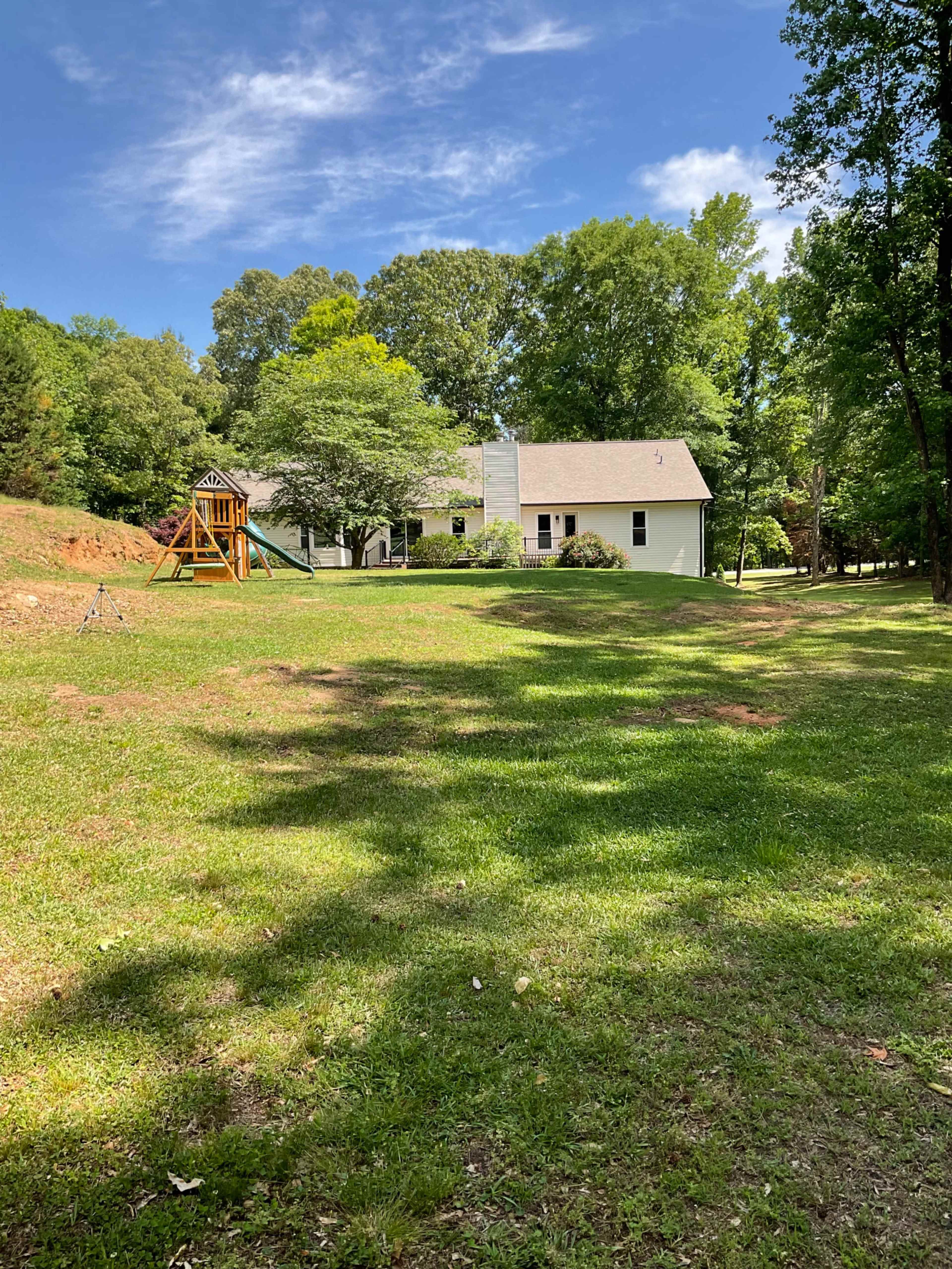 The image shows a spacious backyard with a wooden swing set and a light-colored house set against a backdrop of trees and a blue sky.