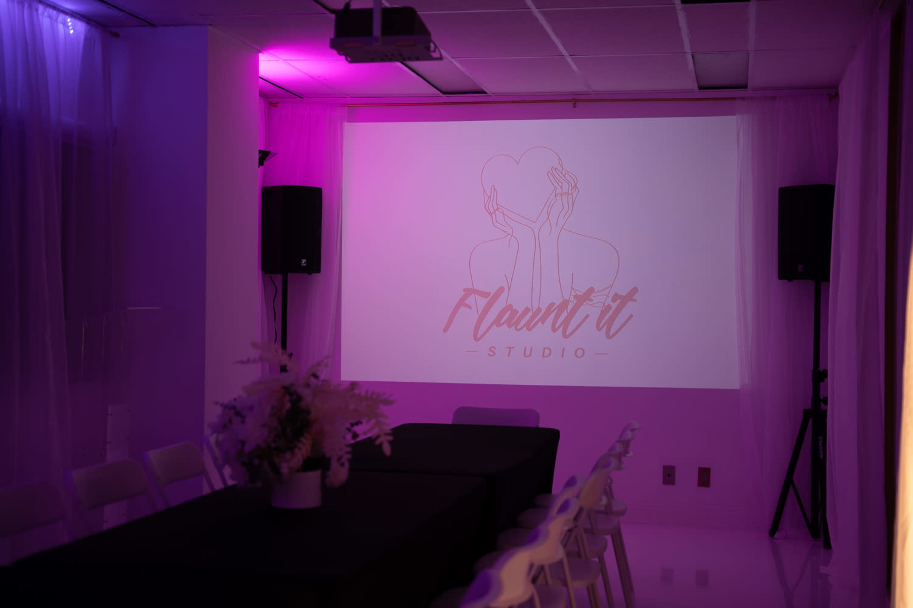 A dimly lit room features a projected logo on a wall, along with a long table and white chairs arranged for an event.